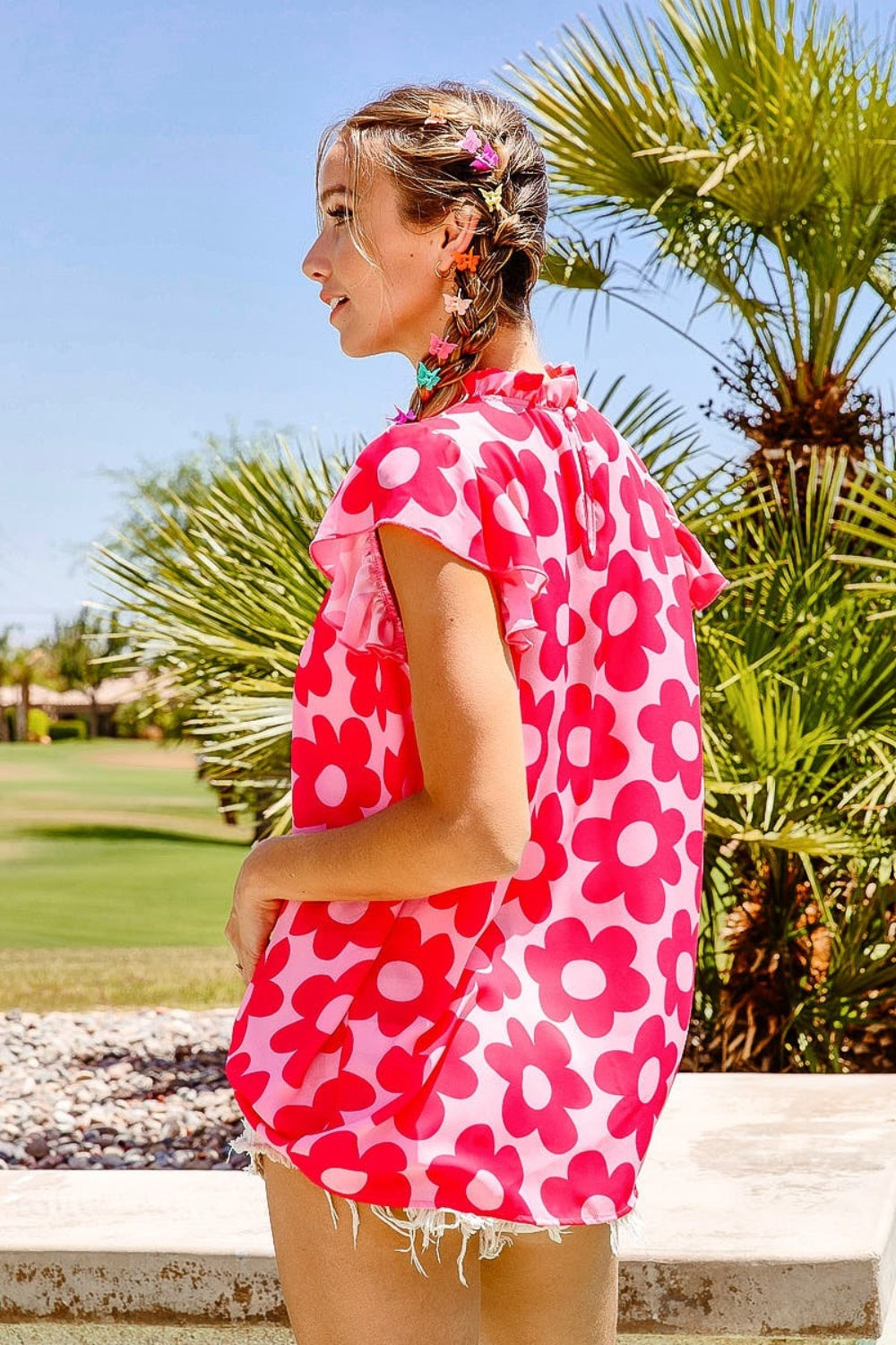 A woman with braided hair wearing a BiBi Floral Contrast Ruffled Top and light denim shorts stands outdoors in front of palm trees on a sunny day.