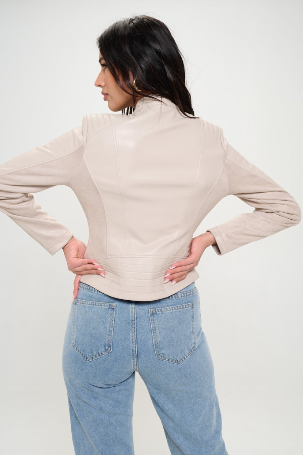 A woman with long dark hair, wearing the cruelty-free Coalition LA Zip Up Vegan Moto Jacket in light beige, pairs it with blue jeans as she stands with her arms crossed.