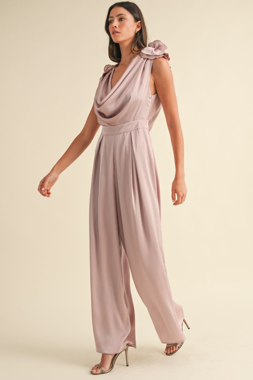 A woman stands against a plain background wearing the MABLE 3D Floral Applique Deep Cowl Neck Jumpsuit, which features an open back and wide-leg pants, paired with open-toed heels.