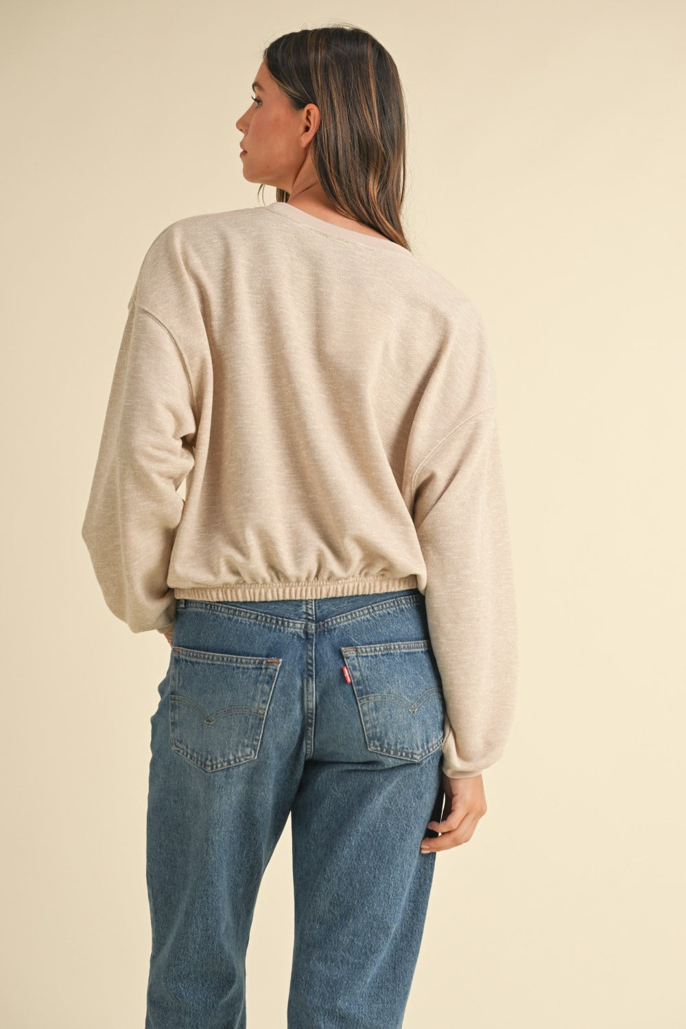 A woman with long brown hair wears the Mable Round Neck Letter Embroidery Crop Sweatshirt in beige, paired with blue jeans, standing confidently with one hand in her pocket against a plain background, perfectly capturing effortless street style.