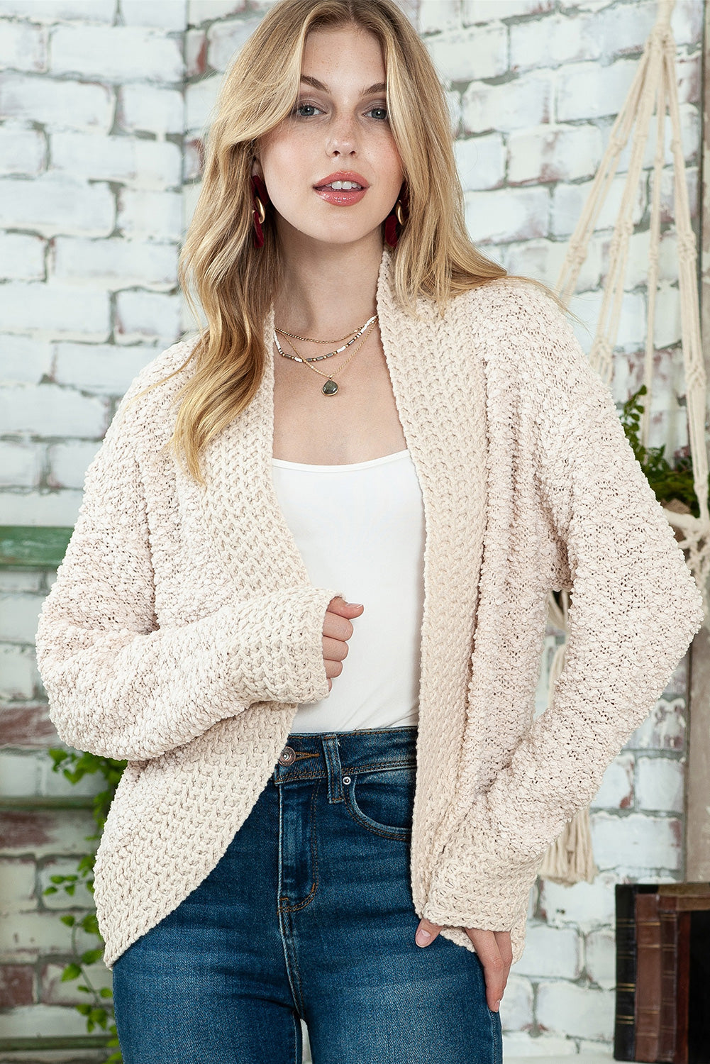 A woman in an Apricot Popcorn Knit Open Front Cardigan and jeans stands against a brick wall, looking to the side with a slight smile.