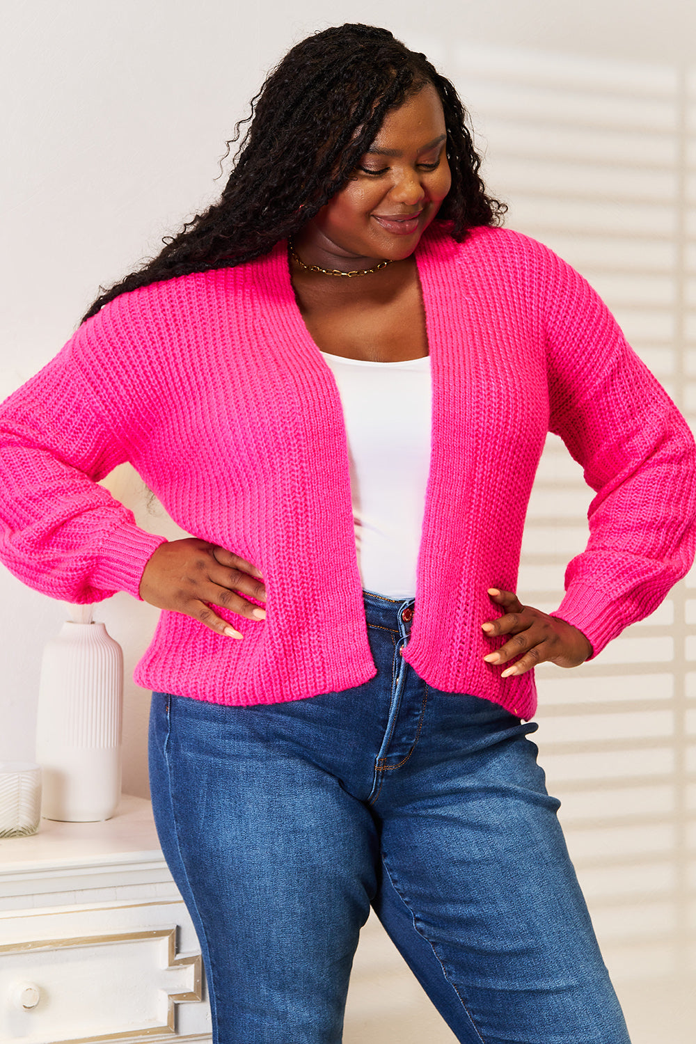 A woman wearing the Woven Right Rib-Knit Open Front Drop Shoulder Cardigan in bright pink over a white top and blue jeans stands indoors, looking to the side.