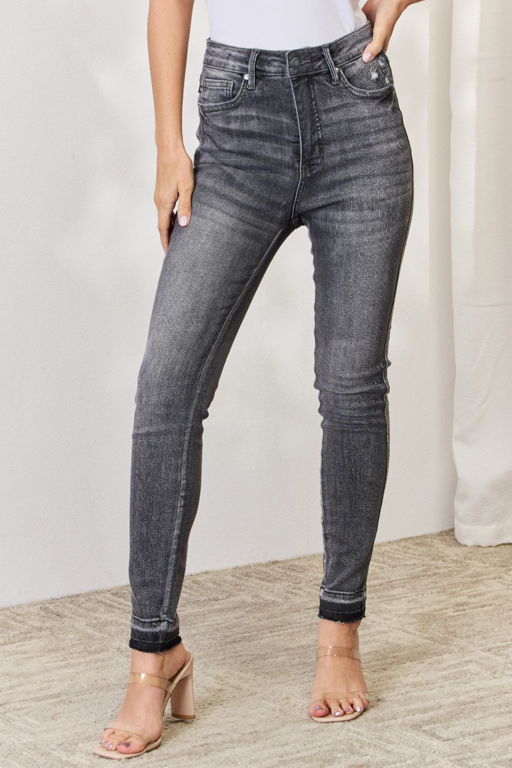 A person wearing the Judy Blue Full Size High Waist Tummy Control Release Hem Skinny Jeans paired with nude heels stands against a light background.