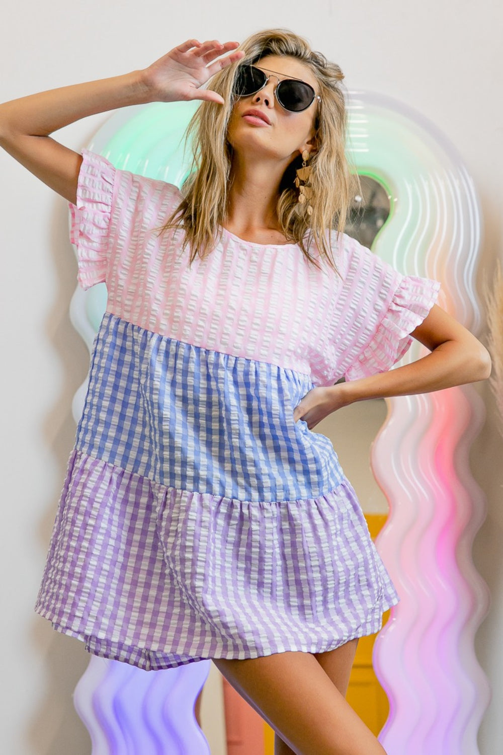 A person in sunglasses and the BiBi Plaid Color Block Tiered Top poses in front of a wavy, colorful mirror.