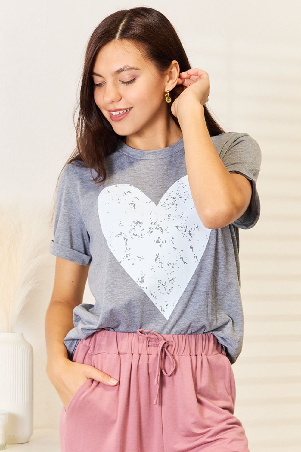 A woman with long hair stands indoors, smiling and touching her hair, wearing the trendy Simply Love Heart Graphic Cuffed Short Sleeve T-Shirt in gray.