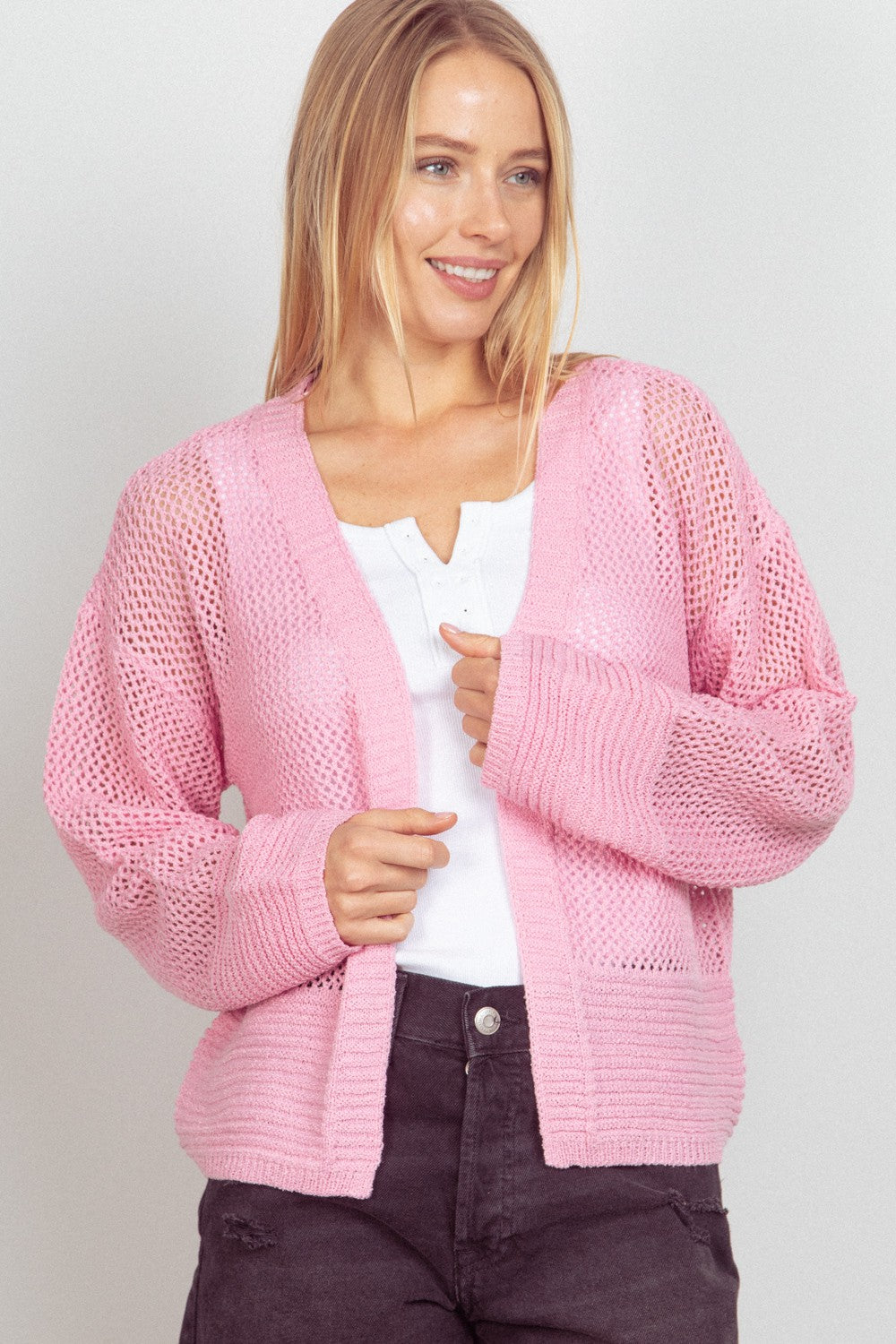 A person wearing a VERY J Eyelet Open Front Long Sleeve Cardigan in pink over a white shirt smiles while standing against a plain background.