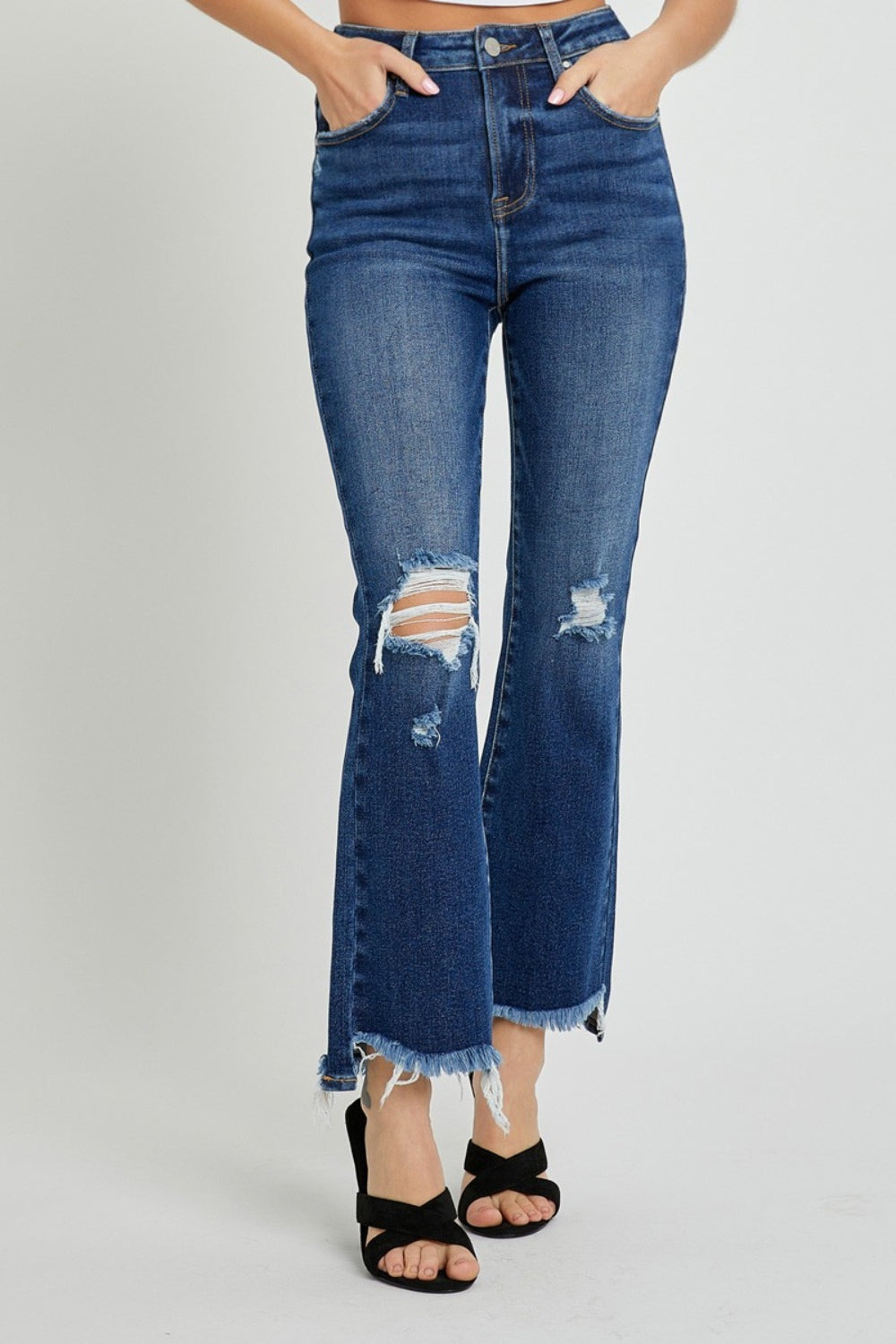 A person wearing RISEN High Rise Distressed Crop Flare Jeans with frayed hems stands with their right hand on their left hip, paired with black open-toed sandals.