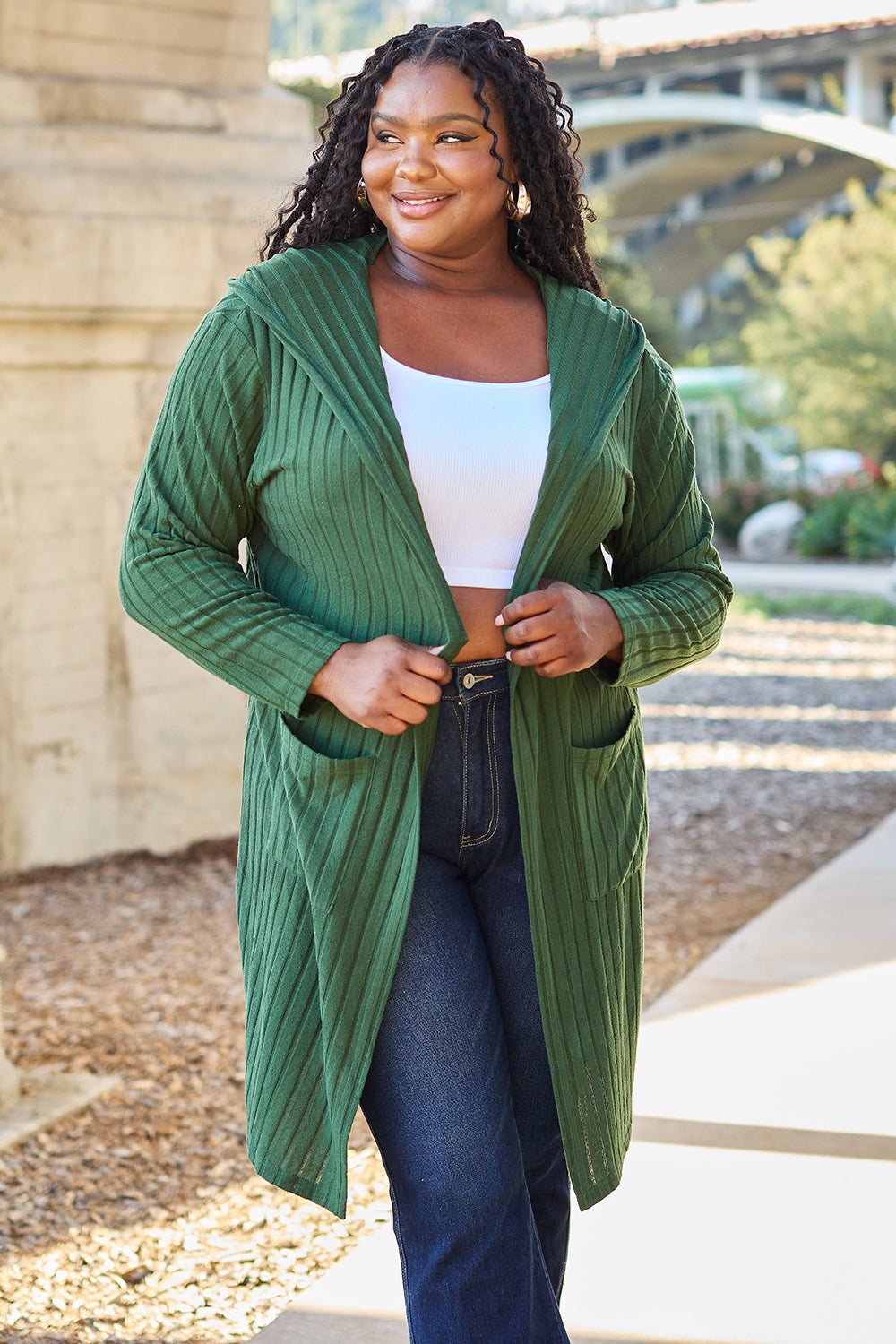 A woman wearing the Basic Bae Full Size Hooded Sweater Cardigan in black, paired with a white top and khaki pants, stands in front of a light background accented by a leafy plant on her left. With one hand tucked into her pocket, she gazes to her right.