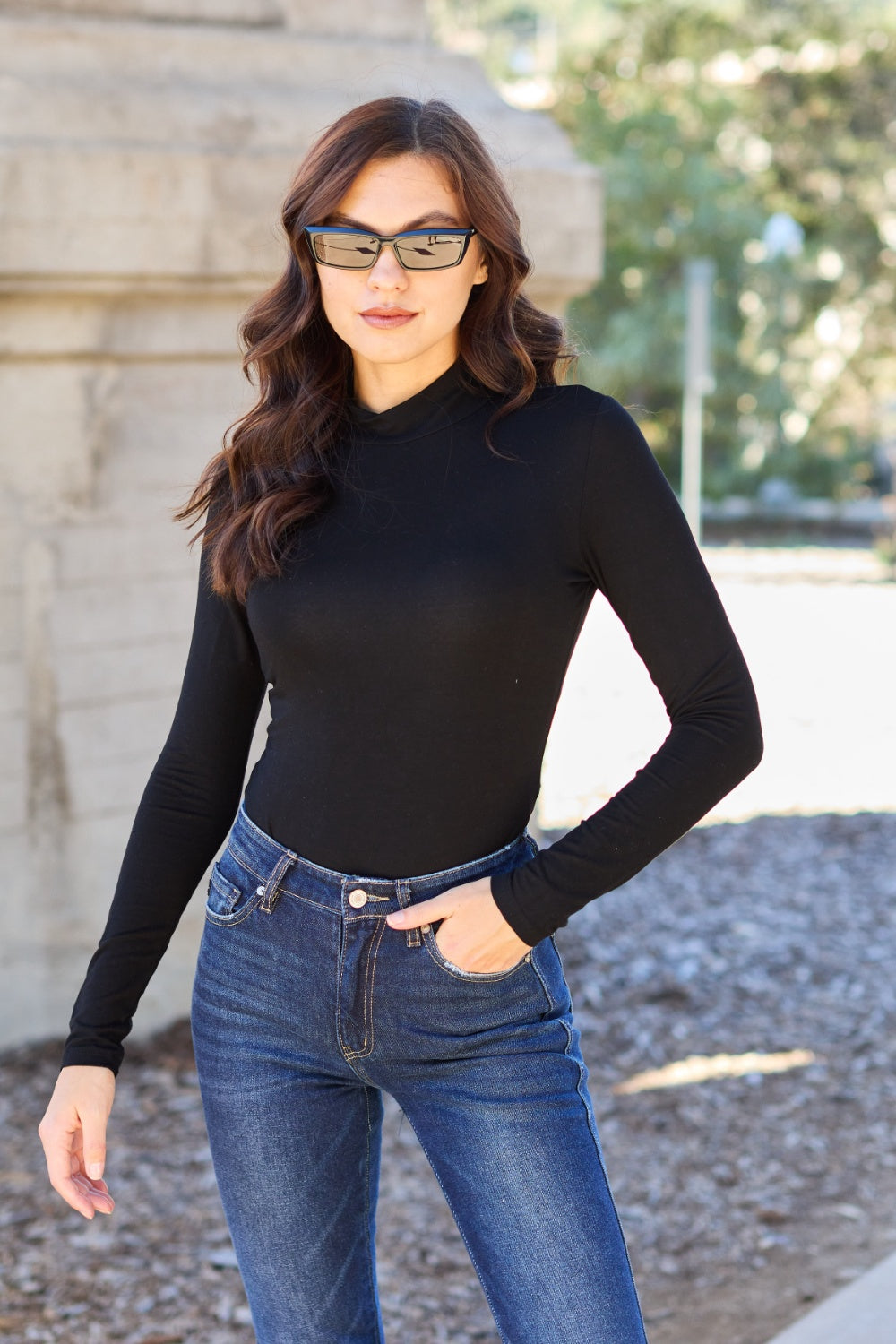 A woman with long, wavy hair stands in front of a light-colored curtain, wearing the Basic Bae Full Size Mock Neck Long Sleeve Bodysuit that offers a basic style and moderate stretch.