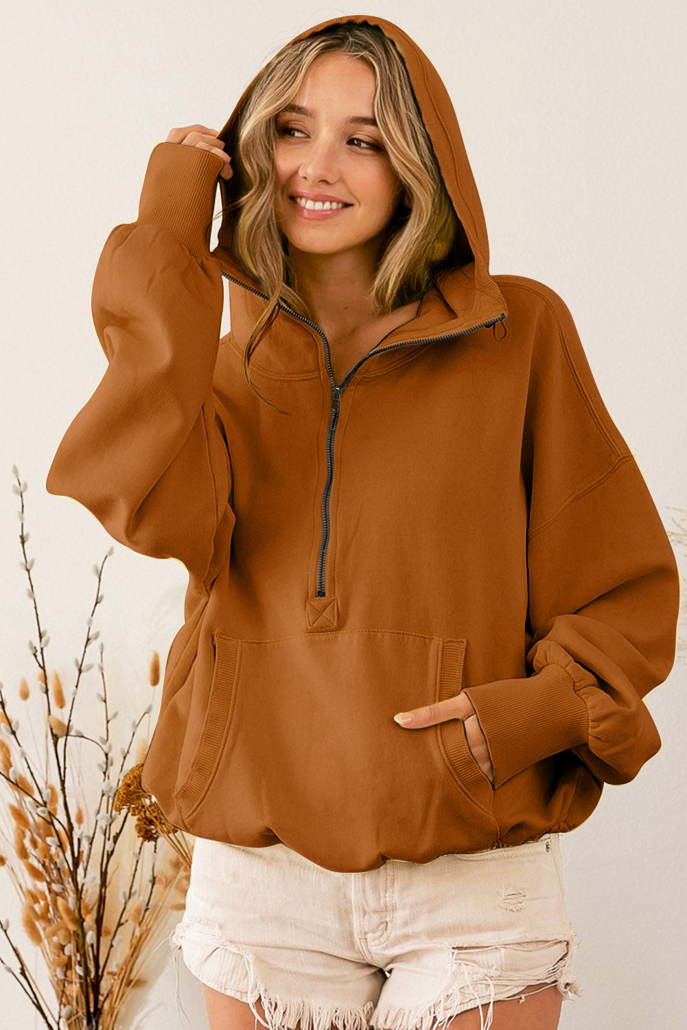 A person wearing a Brown Ribbed Trim Kangaroo Pocket Zipped Hoodie and frayed shorts stands sideways near decorative dried plants in vases.