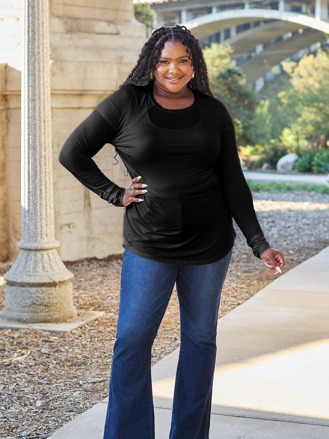 A woman wearing sunglasses and a slightly stretchy Basic Bae Full Size Round Neck Long Sleeve T-Shirt in white stands on a sidewalk with an arched structure in the background, smiling slightly.