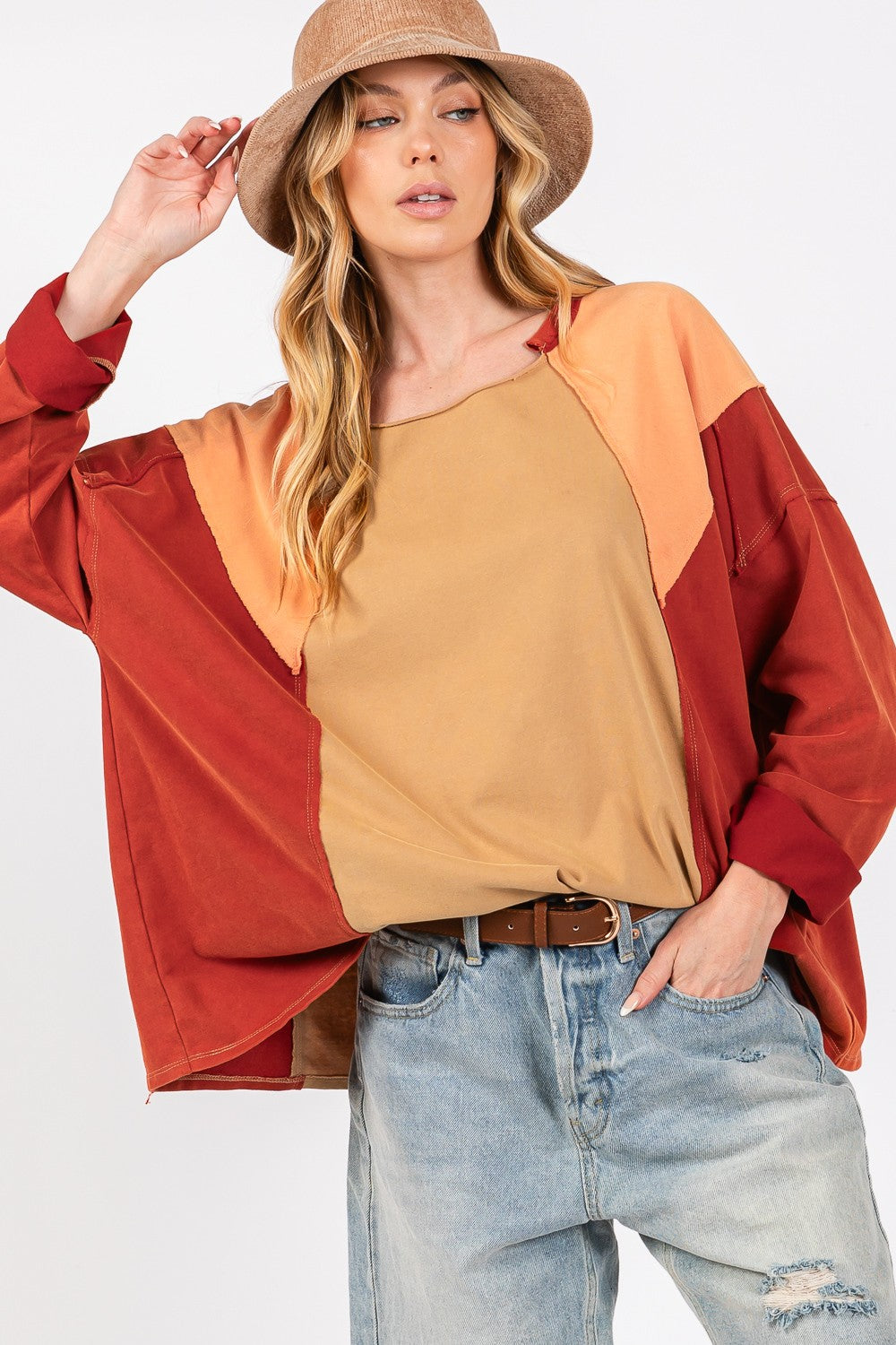 A person in a wide-brimmed hat, wearing the SAGE + FIG Mineral Wash Color Block Top and distressed denim shorts, smiles while adjusting the hat with one hand against a neutral background.