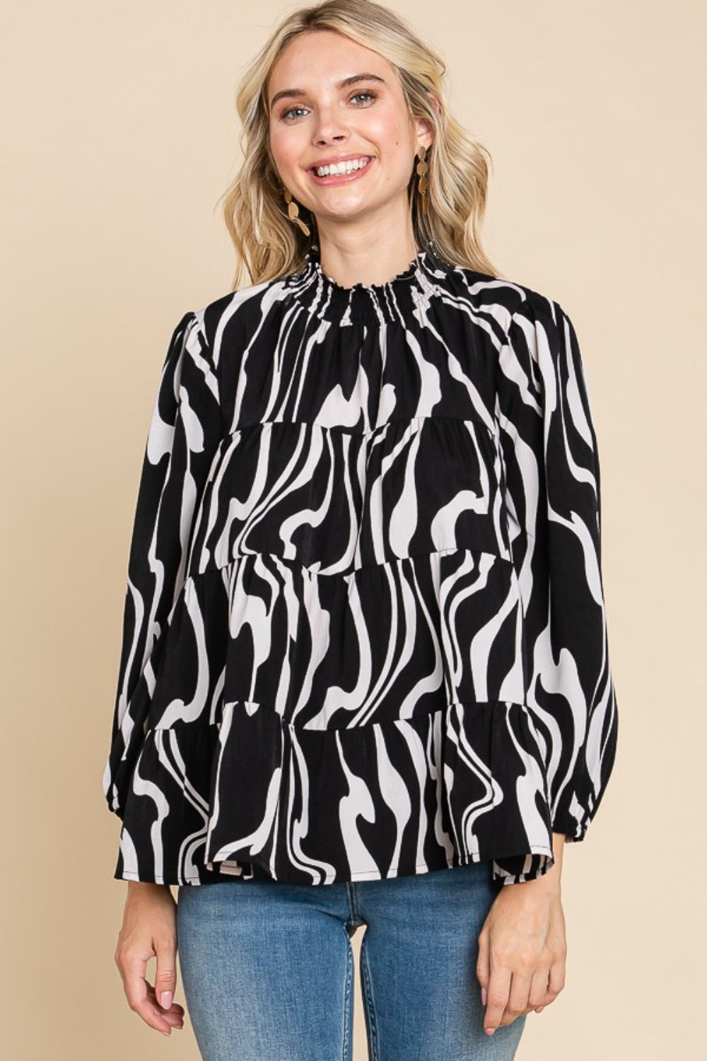 A person wearing a Culture Code Printed Smock Neck Tiered Blouse, featuring a black and white patterned design and long sleeves, paired with blue jeans, stands against a beige background.