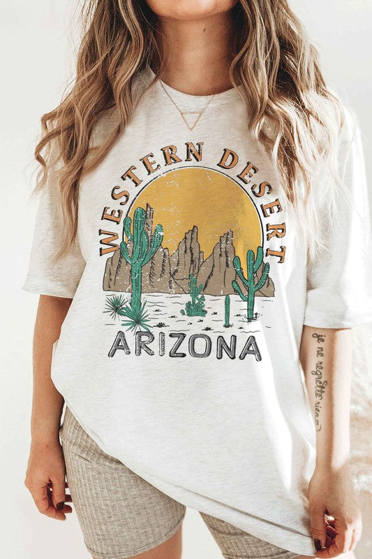 A person wearing the WESTERN DESERT ARIZONA Graphic Tee showcases a bold design featuring cacti and a mountainous landscape against a setting sun. With unisex sizing, this tee is perfect for anyone who loves desert vibes.