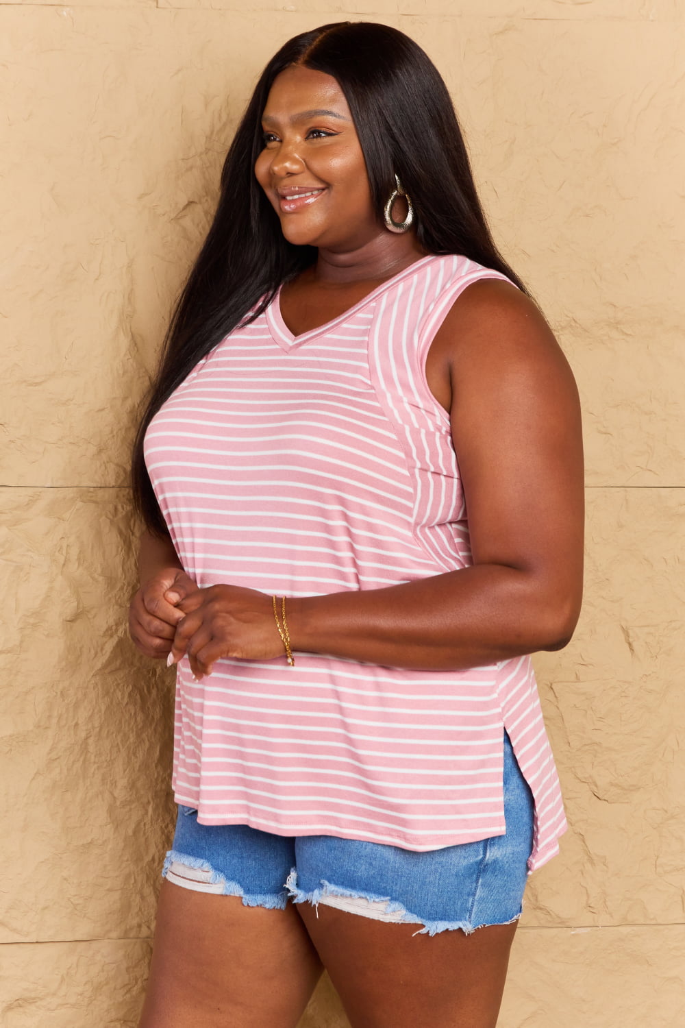 A woman wearing the Doublju Talk To Me Full Size Striped Sleeveless V-Neck Top in pink and white, paired with denim shorts, smiles while standing against a textured beige background.