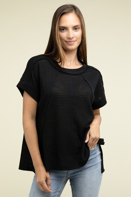 A woman with long brown hair is wearing a purple Brushed Waffle Exposed-Seam Short Sleeve Top and blue jeans, standing against a plain beige background. This casual wardrobe choice, with its short sleeves and side slits, is perfect for everyday wear.