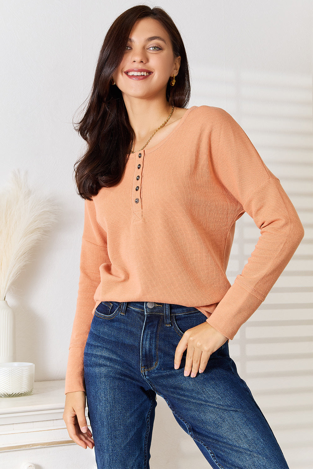 A woman stands indoors, wearing a Basic Bae Half Button Long Sleeve Top in peach and blue jeans, with her hand resting on her hip, showcasing versatile chic wardrobe choices.