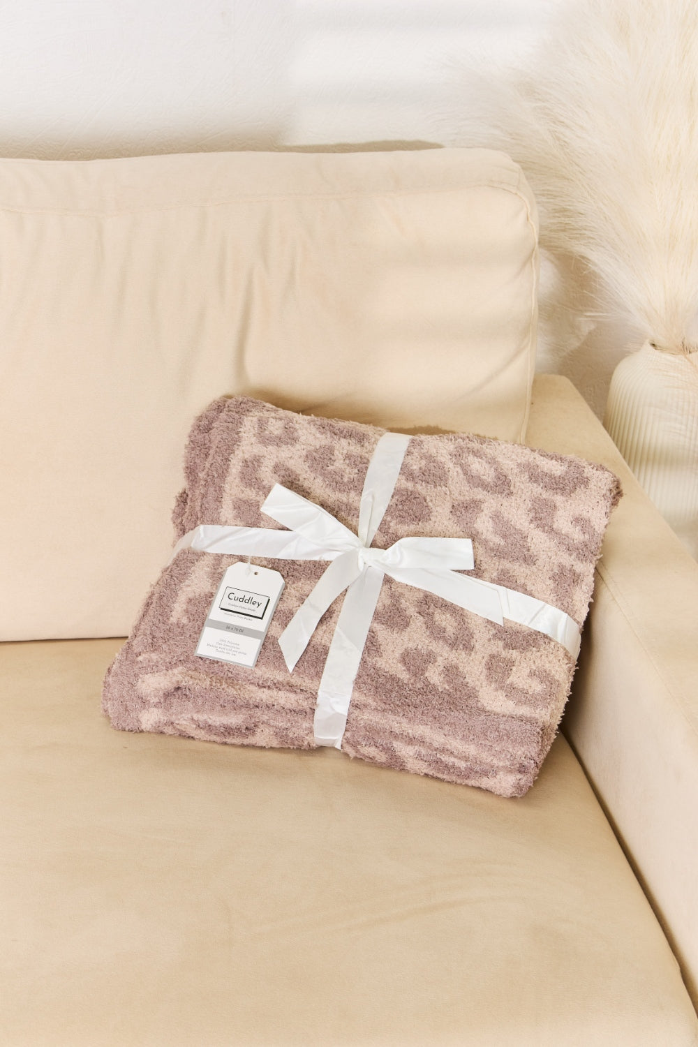 A woman stands in a room holding up the Cuddley Leopard Decorative Throw Blanket, which features a large, leopard-print design in a neutral color scheme. She is smiling and facing the camera, showcasing the 100% polyester fabric.