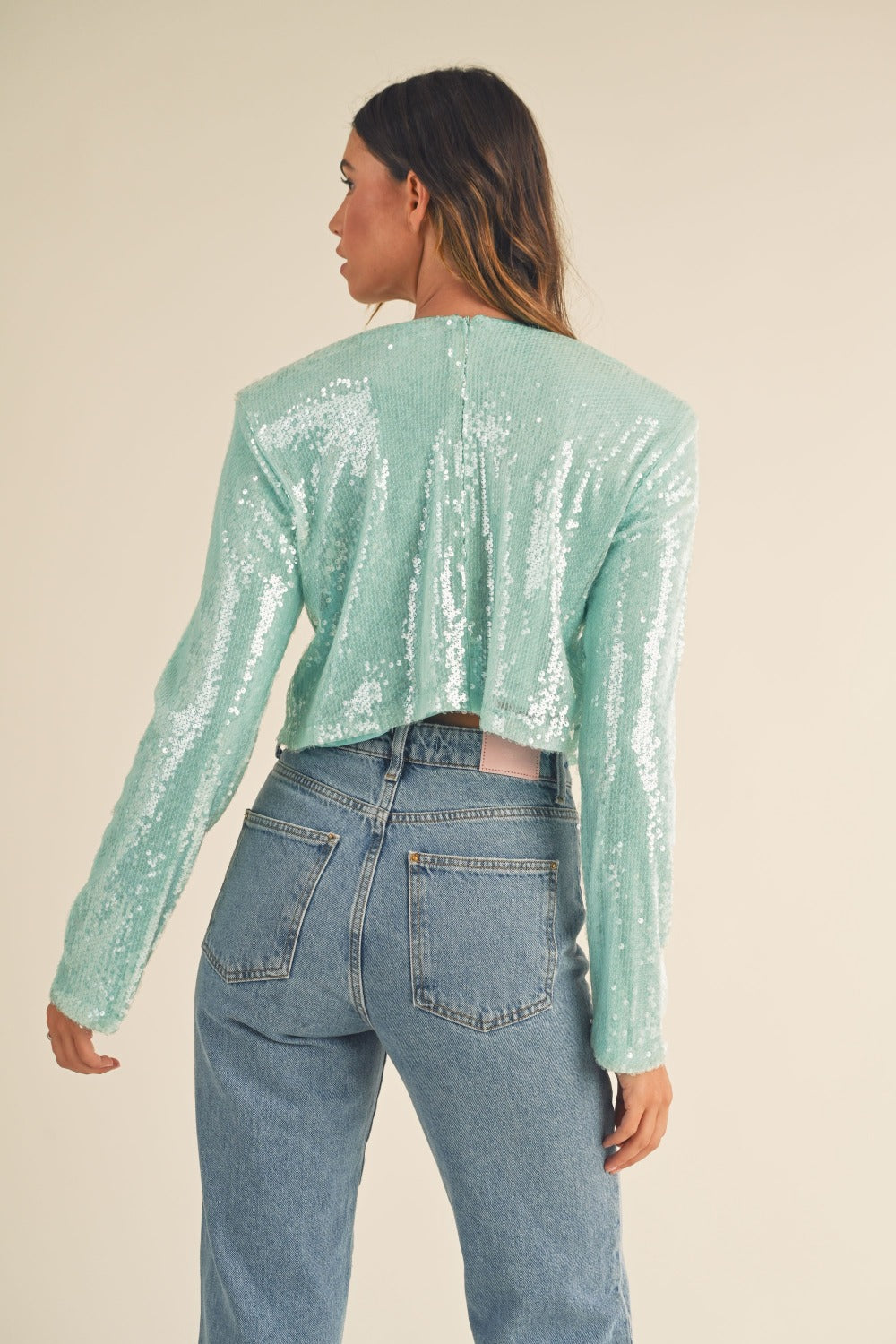 A person with long hair, wearing a fashion-forward, seafoam green MABLE Shoulder Padded Sequin Crop Top and high-waisted blue jeans, stands against a plain background.