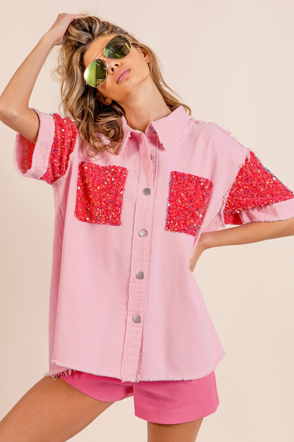 A woman poses in the BiBi Sequin Detail Raw Hem Short Sleeve Shirt, a pink button-up with red sequin detail pockets, paired with matching pink shorts and reflective aviator sunglasses.