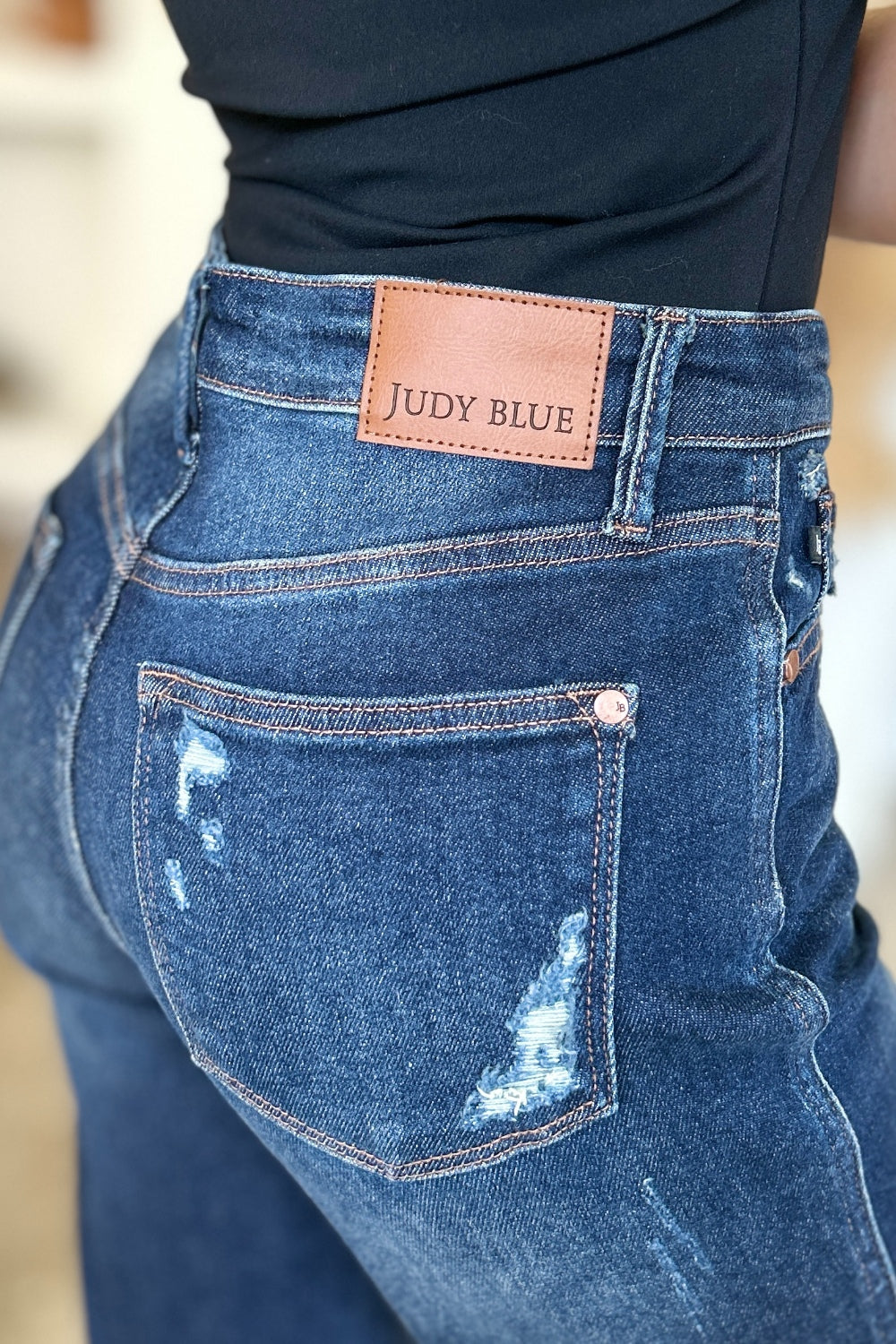 A woman in a black sleeveless top and Judy Blue Full Size High Waist Rigid Magic Heavy Destroy Straight Jeans stands indoors with her hands in her pockets. The background features a plant and shelves adorned with decor items.