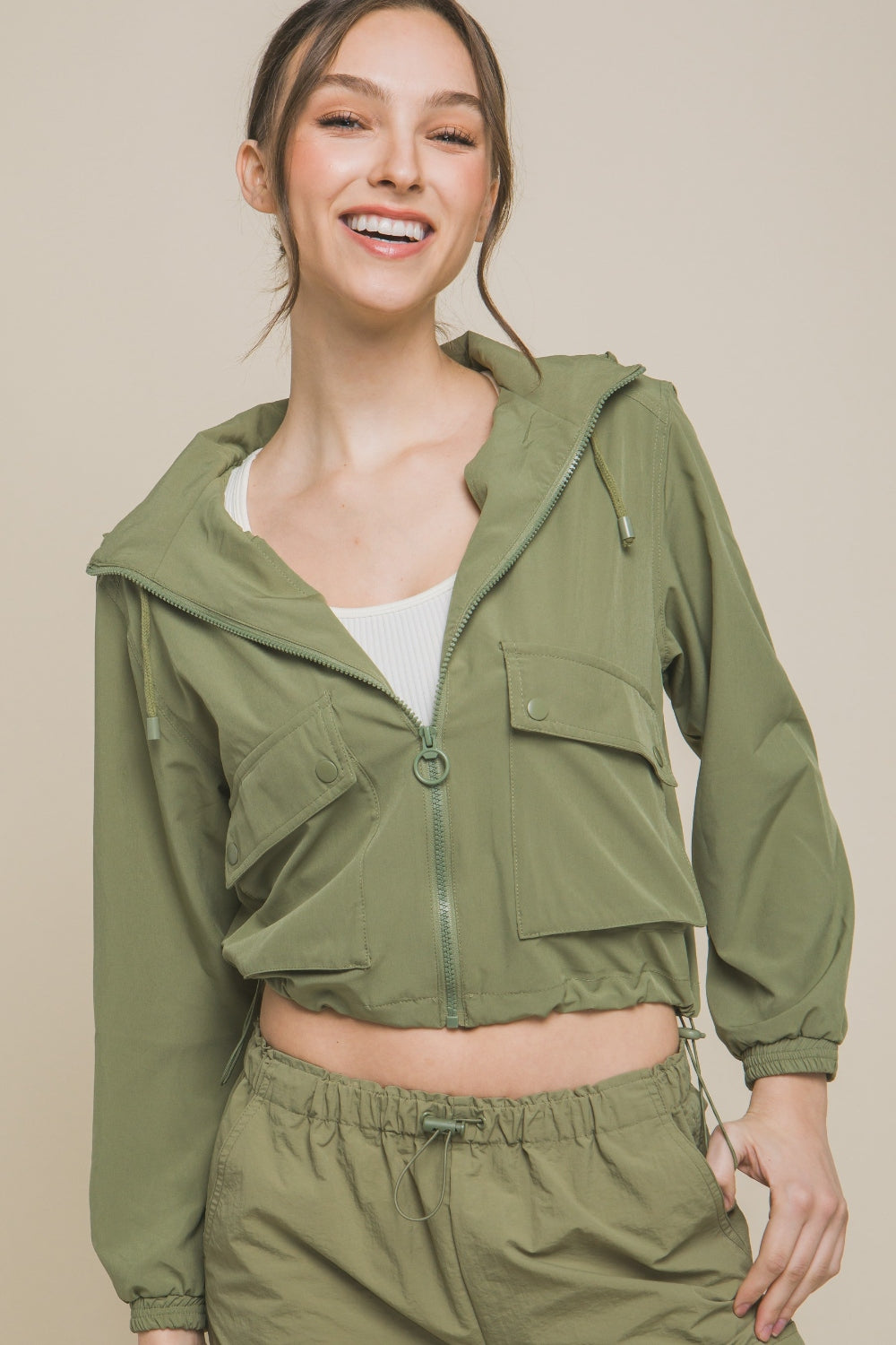 A smiling person is wearing a Love Tree Drawstring Hem Hooded Zip-Up Jacket in light green with matching pants, complemented by a white top underneath, and is standing against a plain background.