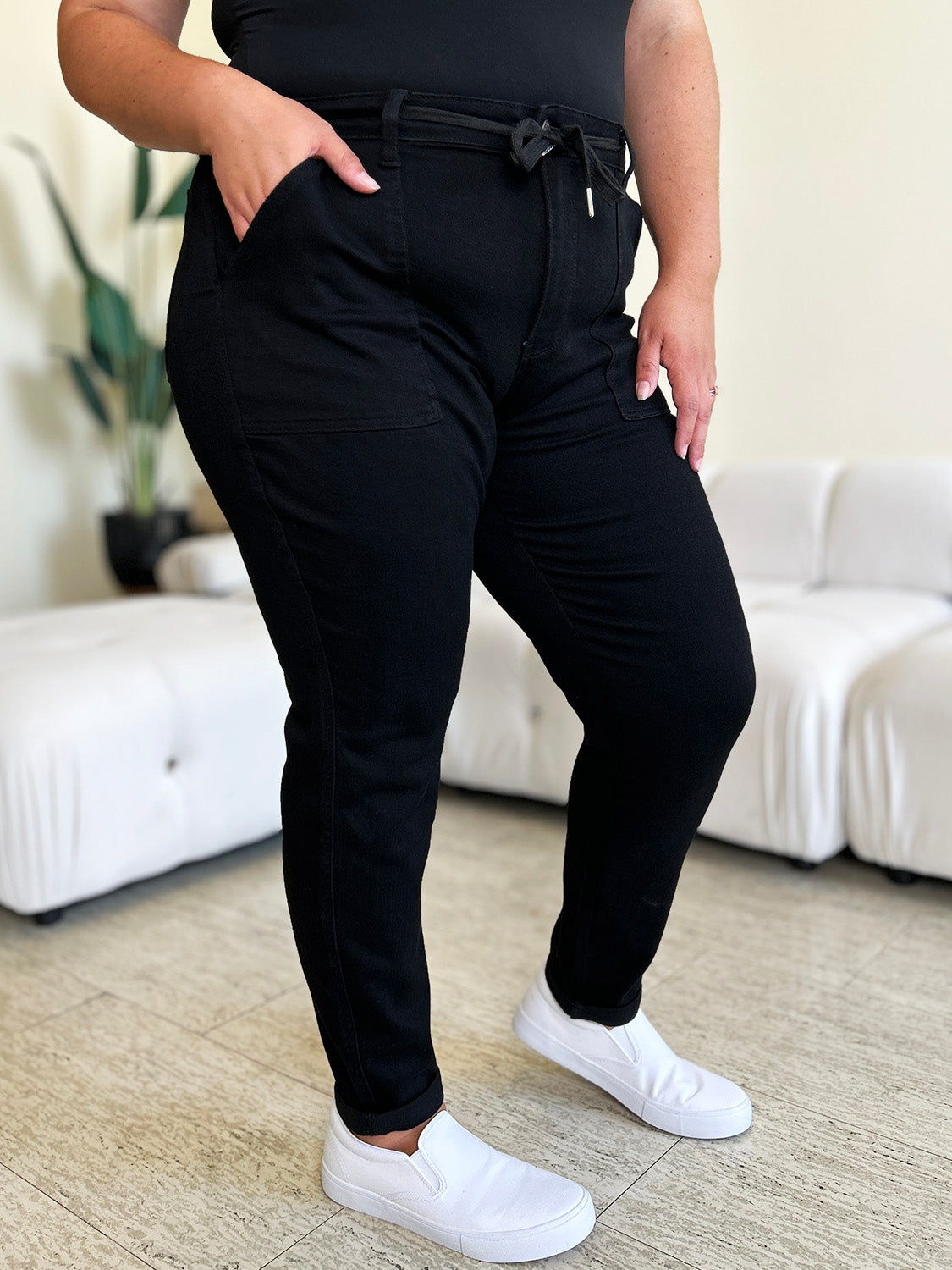 Person standing indoors, wearing a white top, black Judy Blue Full Size High Waist Double Roll Cuff Jeans, and white slip-on shoes, next to white couches and a plant in the background.