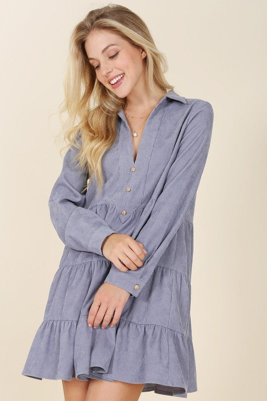 The woman in the Corduroy tiered dress, featuring a deep V-neck, smiles as she gently touches her forehead, showcasing the light blue fabric cascading elegantly.