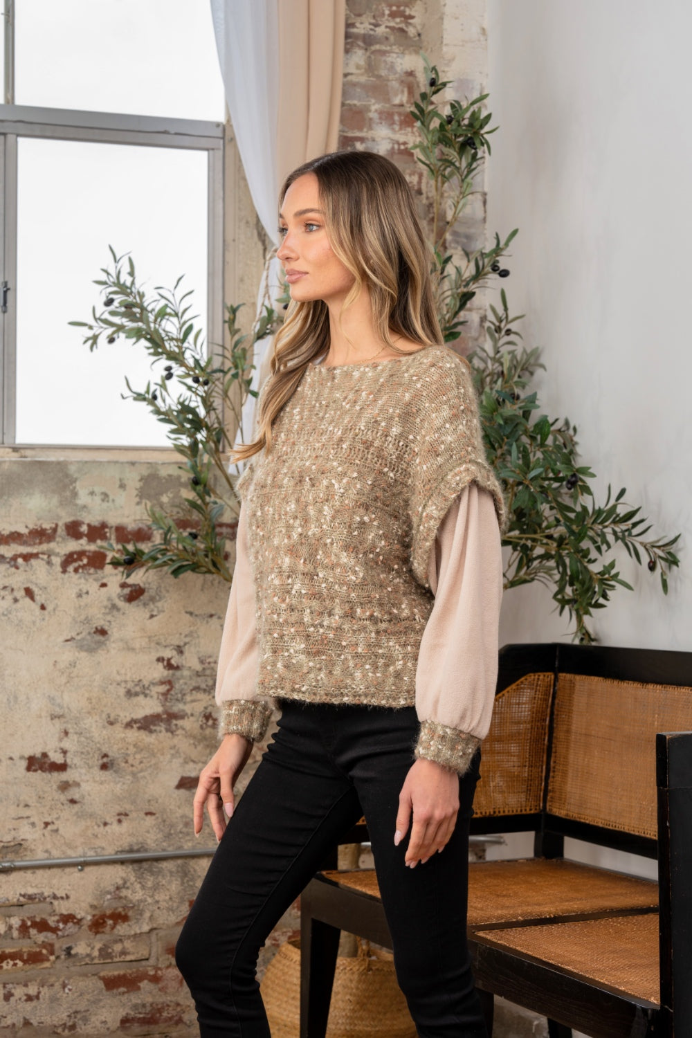 Standing indoors by a window, a person looks cozy and stylish in black pants, their textured sweater contrasting perfectly with the brick wall and greenery backdrop. The Sew In Love Full Size Fuzzy Long Sleeve Knit Top adds an elegant touch as one hand rests in their pocket.