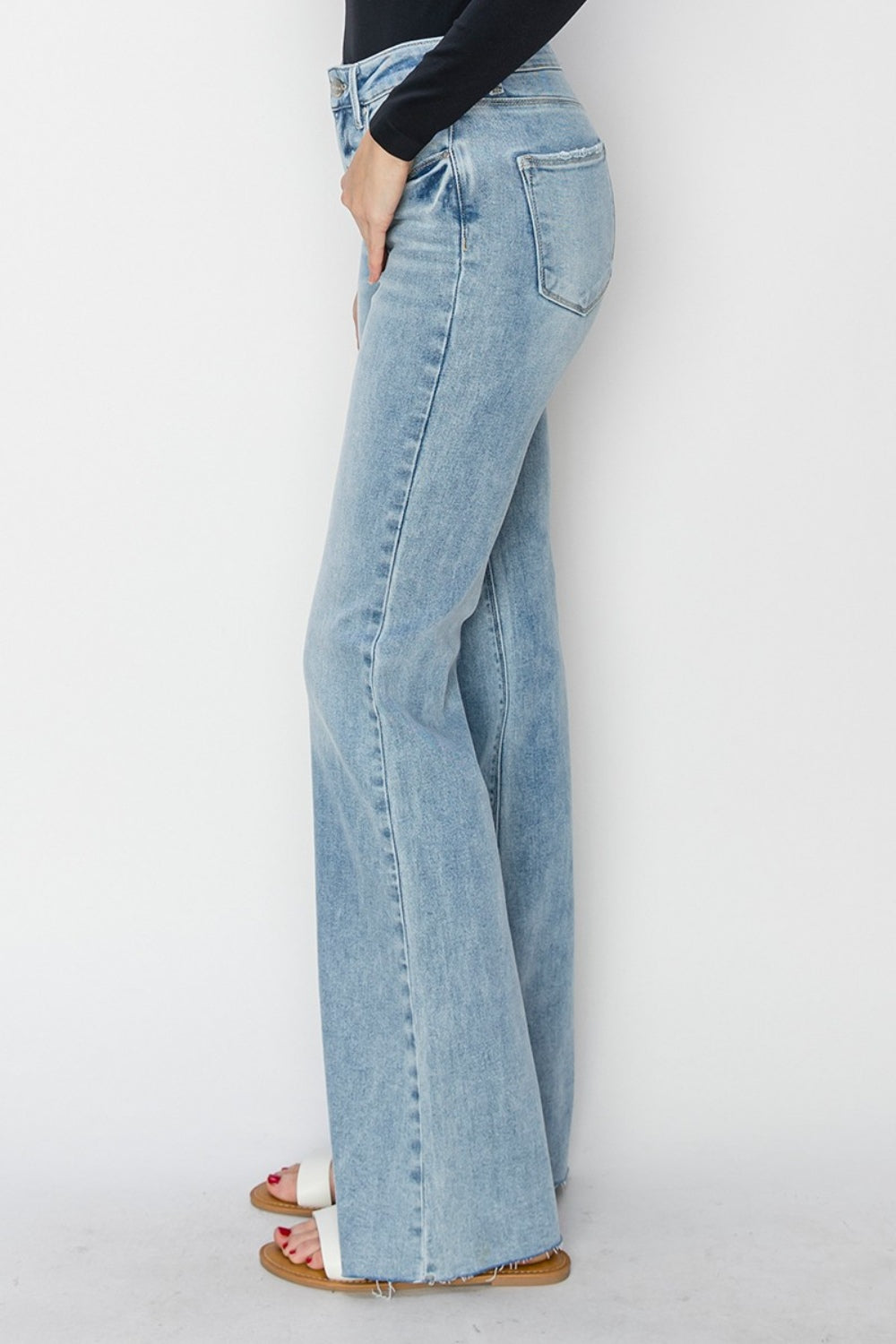 A person wearing RISEN Full Size High Rise Raw Cut Hem Bootcut Jeans in a light wash and white sandals, standing against a plain white background.