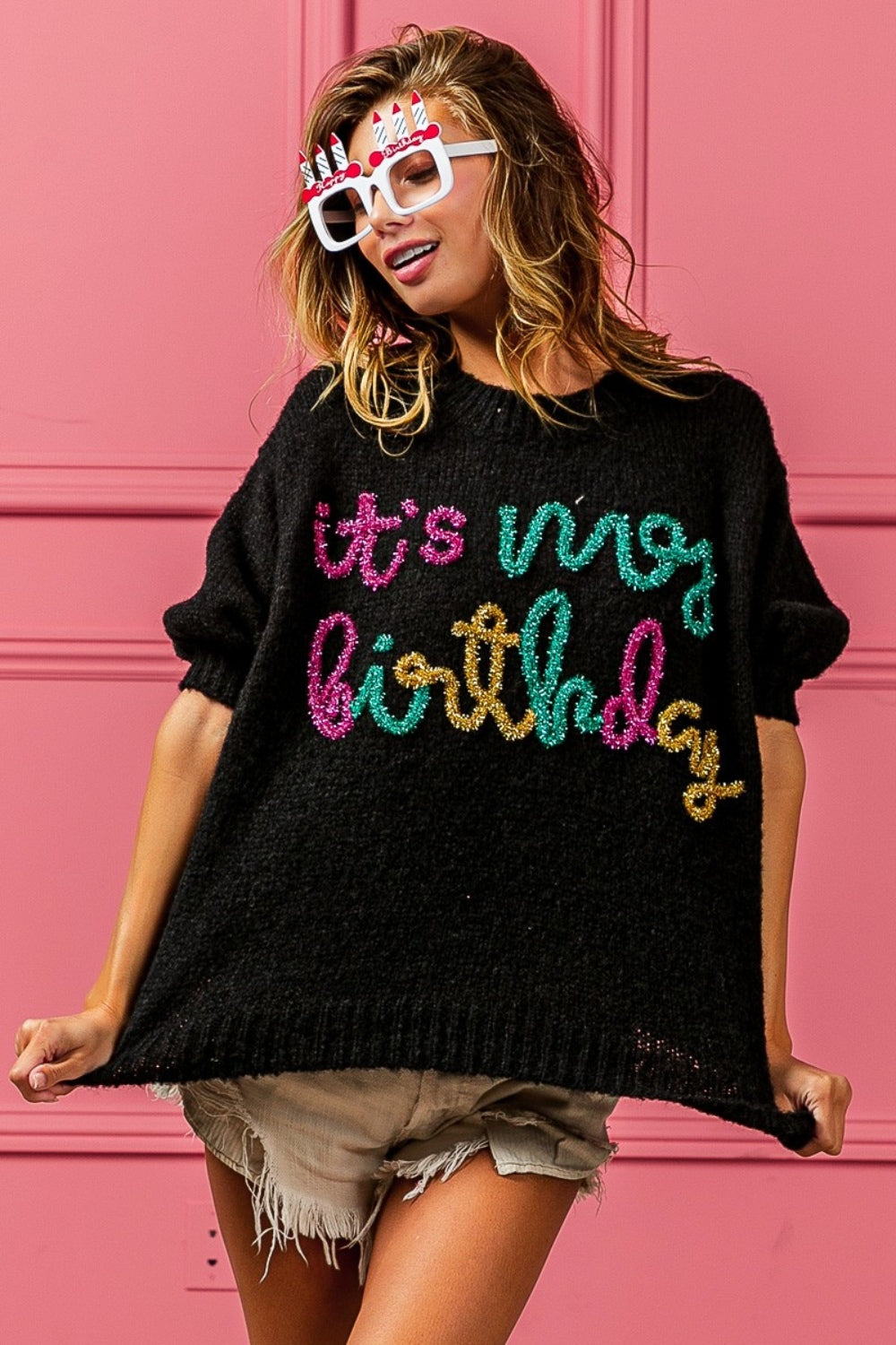 A woman poses against a pink wall, wearing a BiBi Metallic Letter Puff Sleeve Hairy Sweater that reads "It's My Birthday," beige shorts, and novelty glasses with birthday candles.