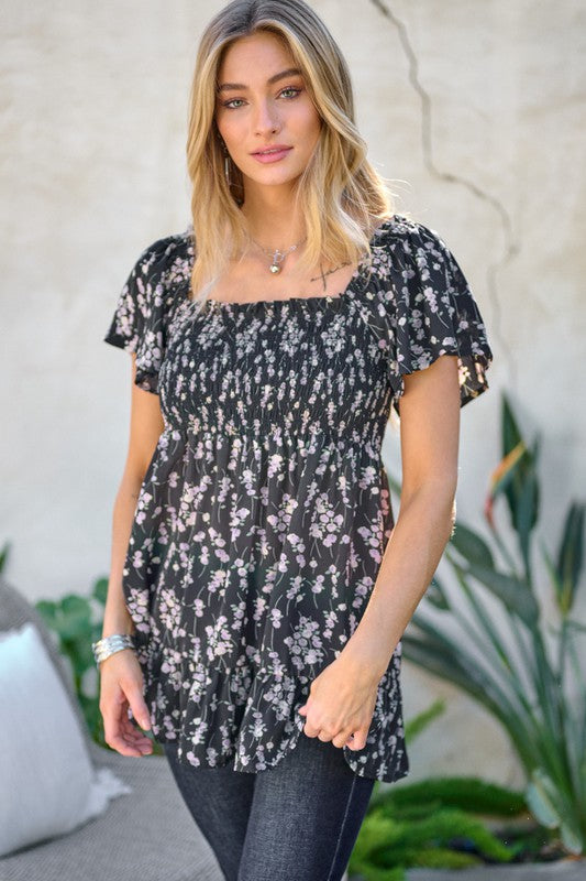 A woman stands outdoors donning a Floral Printed V-Neck Ruffle Top paired with jeans, set against a neutral background, with plants in the foreground, highlighting her fashion-forward wardrobe.