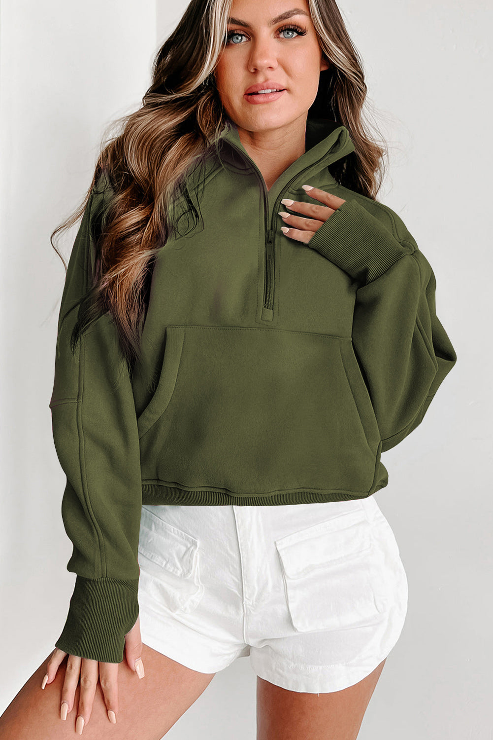 A person with long, wavy hair stands in front of a white and black background, wearing the Green Fleece Lined Zip Up Stand Collar Thumbhole Sleeve Sweatshirt paired with white shorts, creating a stylish look perfect for casual outings.