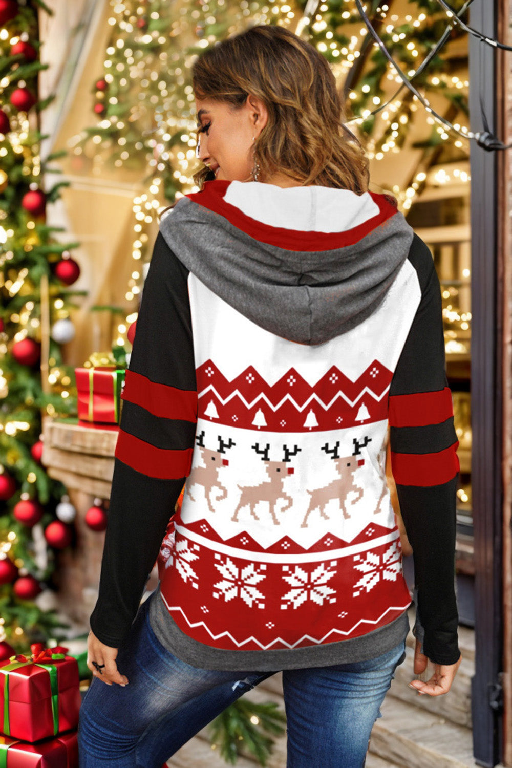 A person is wearing the Double Take Full Size Christmas Drawstring Long Sleeve Hoodie, which features a holiday-themed red and white pattern with reindeer and snowflakes. This cozy hoodie, made from a polyester spandex blend, has stylish black sleeves accented with red stripes. The person is smiling and looking to the side.