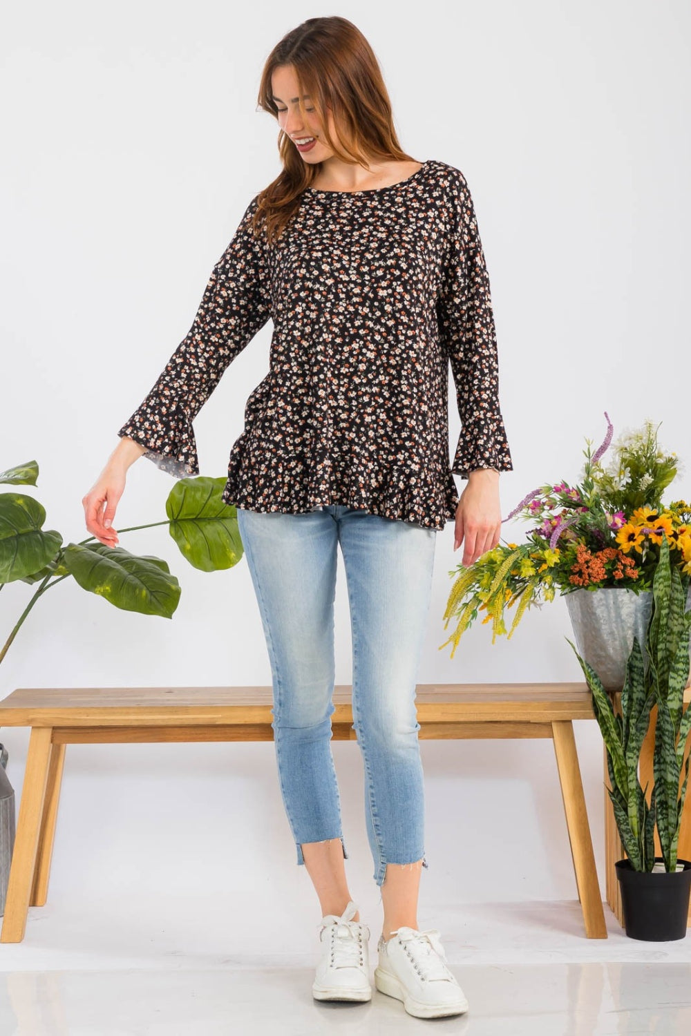 A woman elegantly stands beside colorful flowers against a plain background, embodying feminine fashion in the Celeste Full Size Floral Ruffle Detail Top, which features a round neck and floral patterns, paired with light blue jeans.