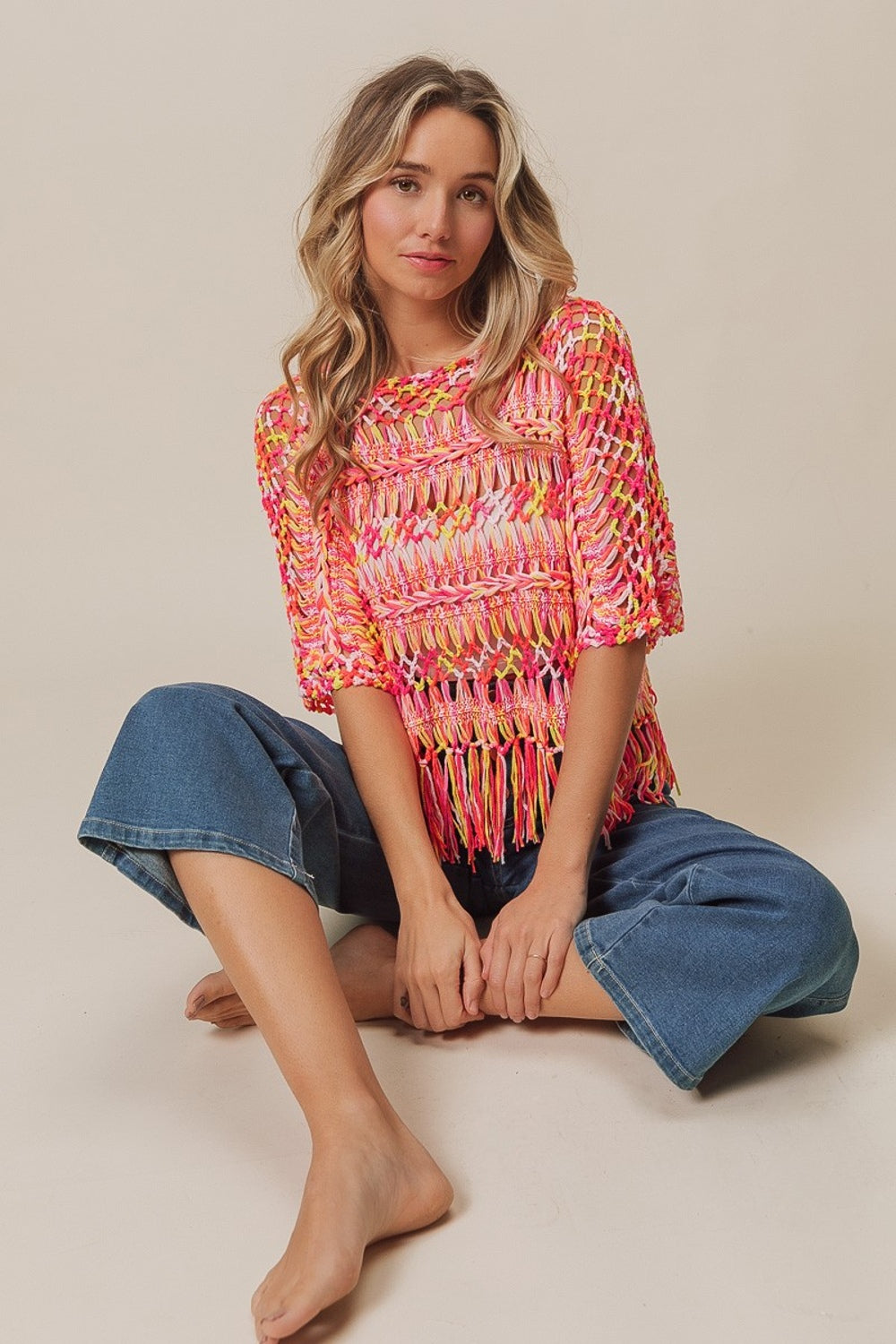 A person with long hair wears the BiBi Openwork Fringed Knit Cover Up, exuding bohemian flair, paired with jeans. They sit with one hand raised and the other resting on their lap against a plain background.