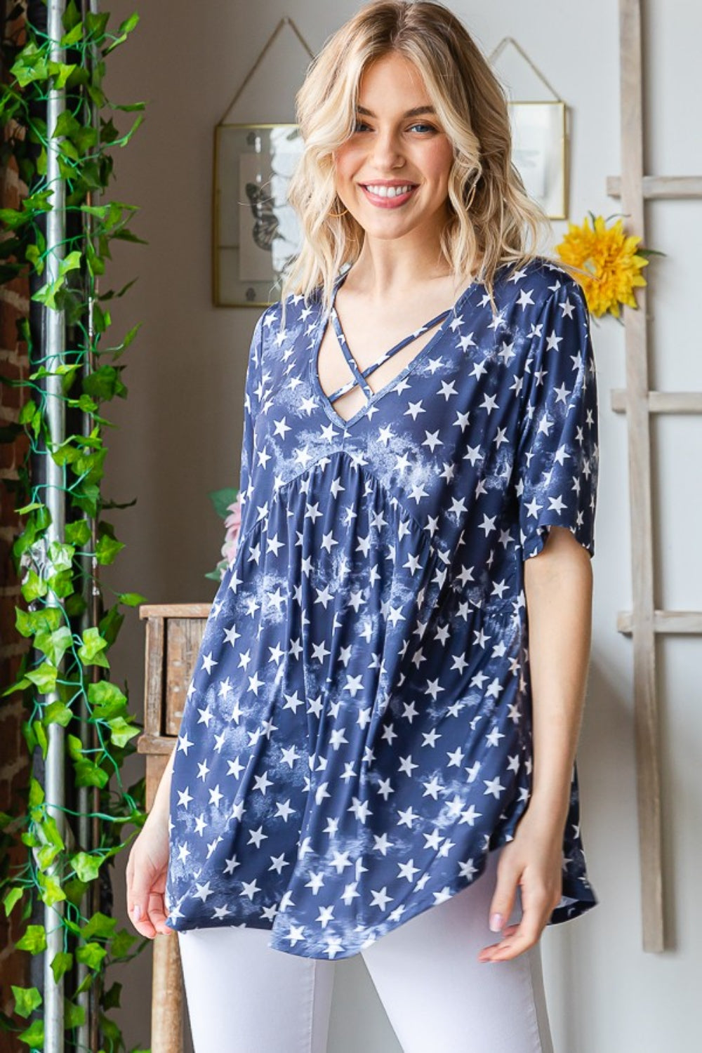 A person with long blonde hair is wearing the Heimish Full Size Short Sleeve Crisscross Star Babydoll Top in blue and white pants, standing indoors near a table adorned with flowers and framed pictures on the wall.