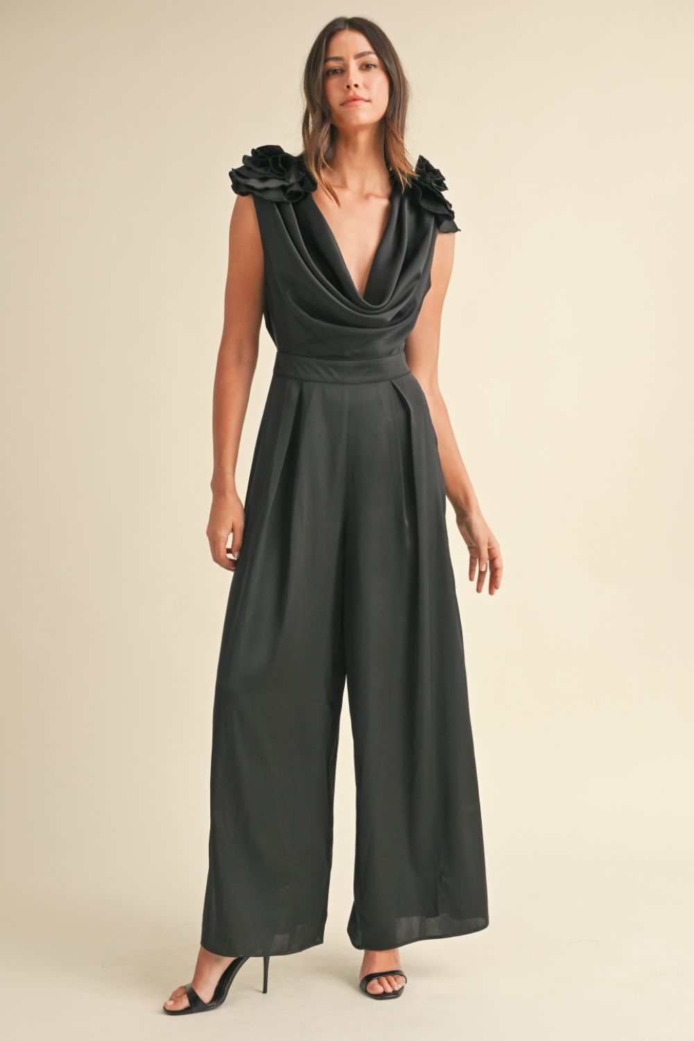 A woman stands against a plain background wearing the MABLE 3D Floral Applique Deep Cowl Neck Jumpsuit in black satin, paired with black high-heeled sandals.