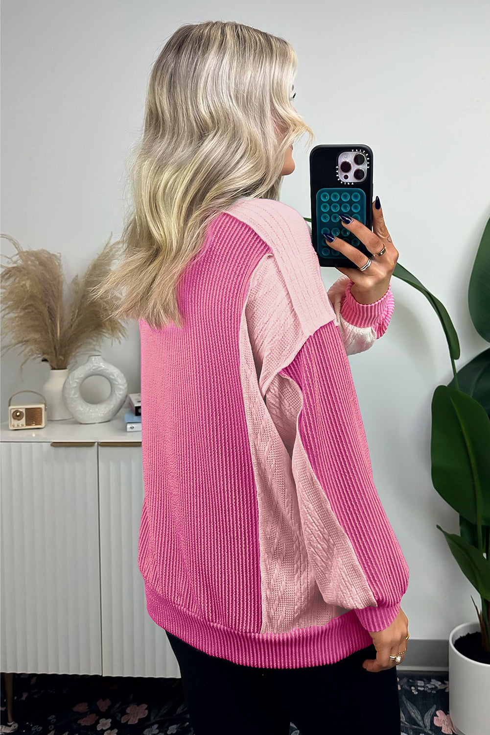 A person with long, wavy blonde hair is taking a selfie in a mirror, wearing the Bonbon Cable Knit Colorblock Exposed Seam Sweatshirt. A plant and decor items are visible in the background.
