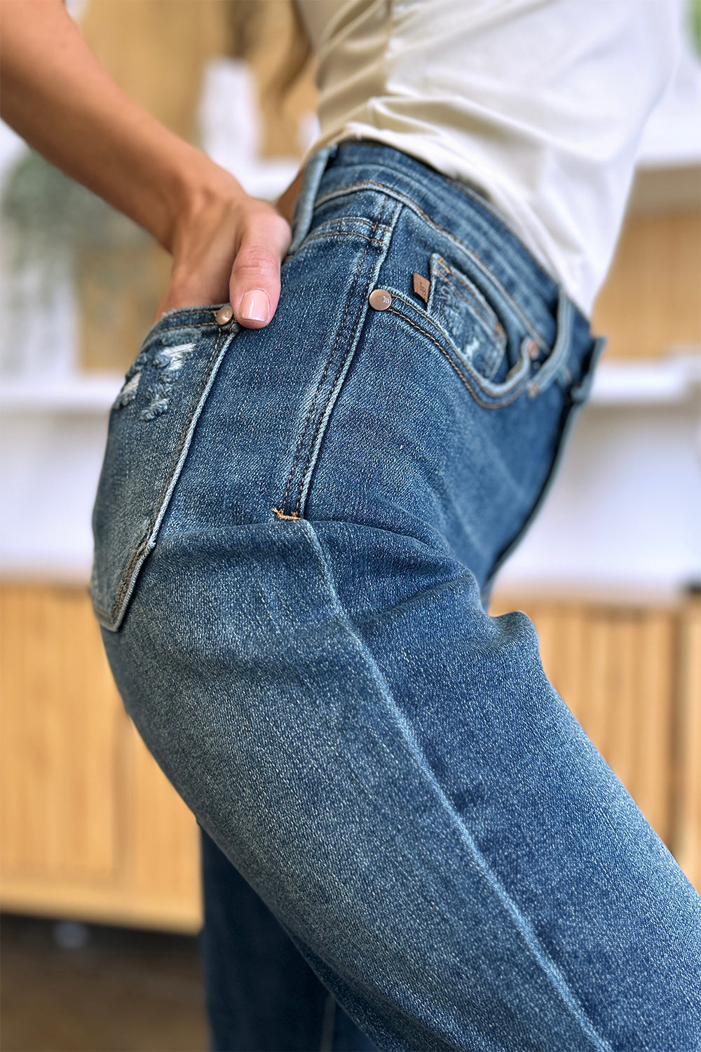 Person wearing Judy Blue Full Size Tummy Control High Waist Slim Jeans in blue and a white shirt, with their hands in their pockets. They are standing indoors on a polished concrete floor with a wooden cabinet and plant in the background.