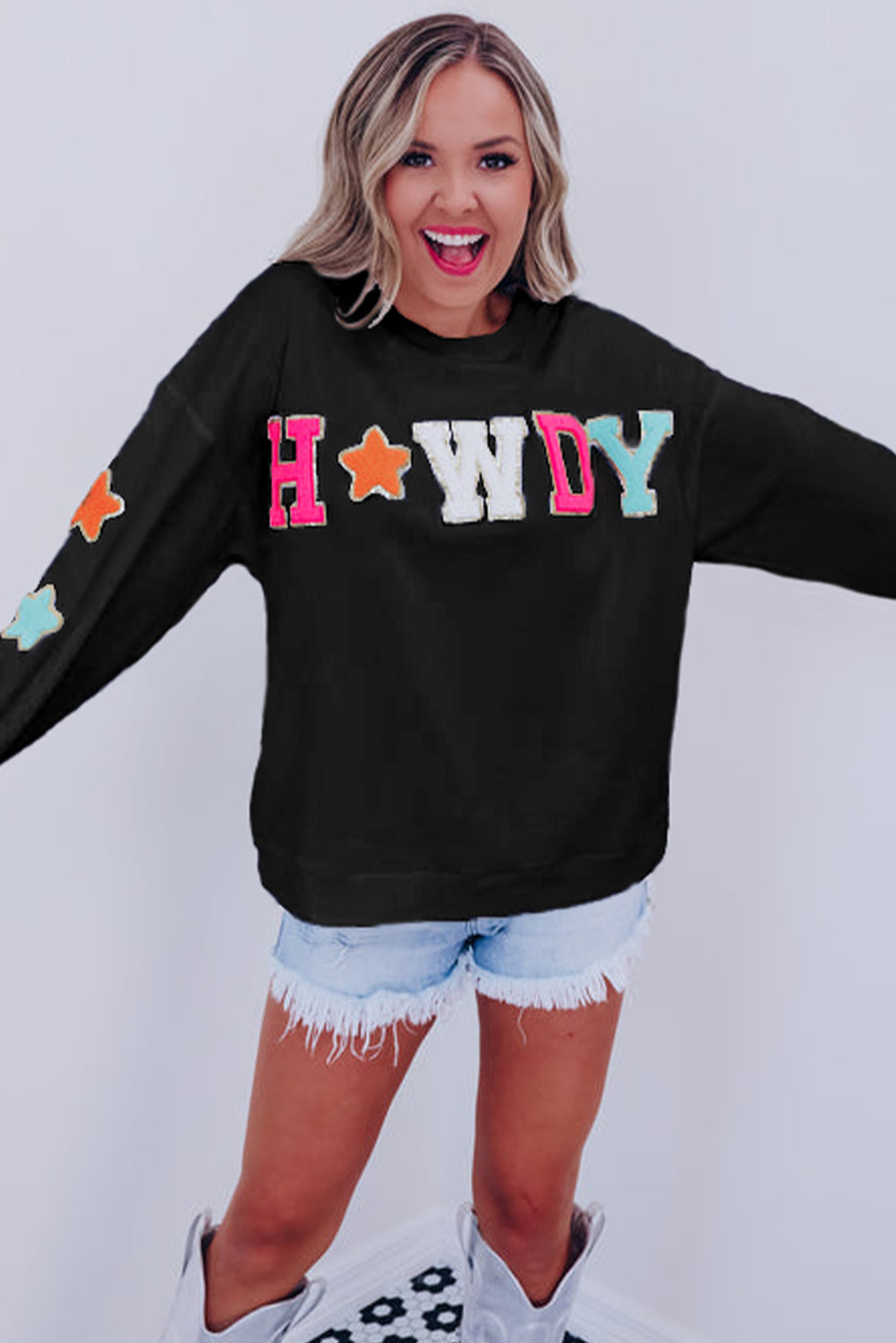 A woman with short blonde hair is wearing the Black Howdy Glitter Chenille Patch Graphic Casual Sweatshirt, featuring colorful star patterns and "WD" letters. She pairs it with light denim shorts and smiles while looking to the side.