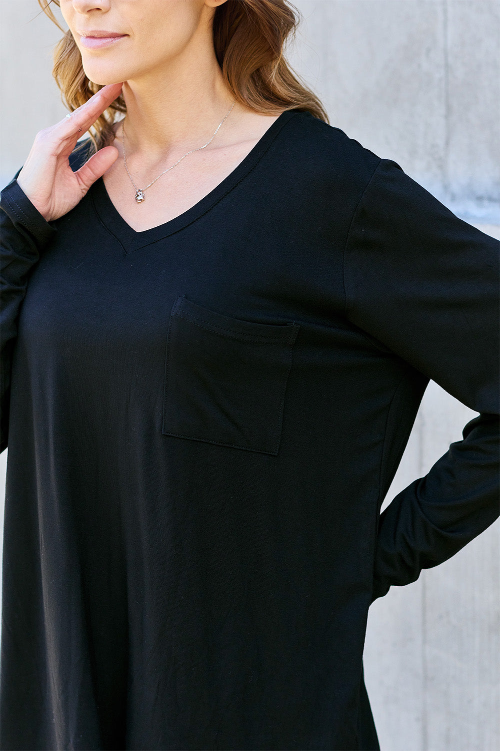 A woman with light brown hair stands against a concrete wall, wearing the Basic Bae Full Size V-Neck Long Sleeve Top in olive green and blue jeans, with a white backpack on her shoulder. Her outfit exudes basic style and is made from polyester, ensuring it’s both comfortable and suitable for machine wash cold. She is looking at the camera.