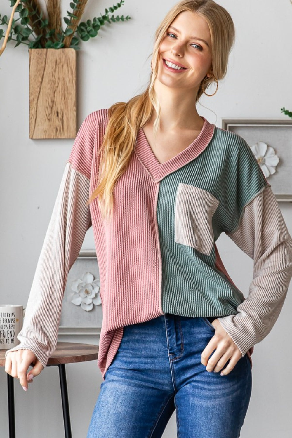 A person with long blonde hair stands indoors, smiling. They are wearing the Heimish Color Block Exposed Seam Ribbed T-Shirt, featuring pink, green, and beige sections, paired with blue jeans. Decorative wall pieces hang in the background.