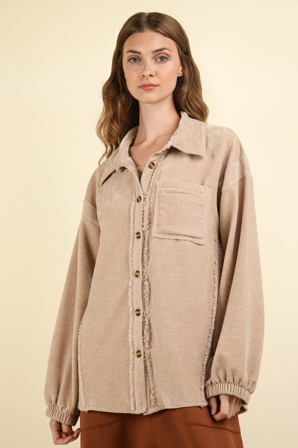 A woman with long wavy hair wears the VERY J Mixed Media Button Down Raw Hem Shacket featuring a drop shoulder design over a brown outfit. She faces the camera with one hand holding the lapel of her shacket against a plain beige background.