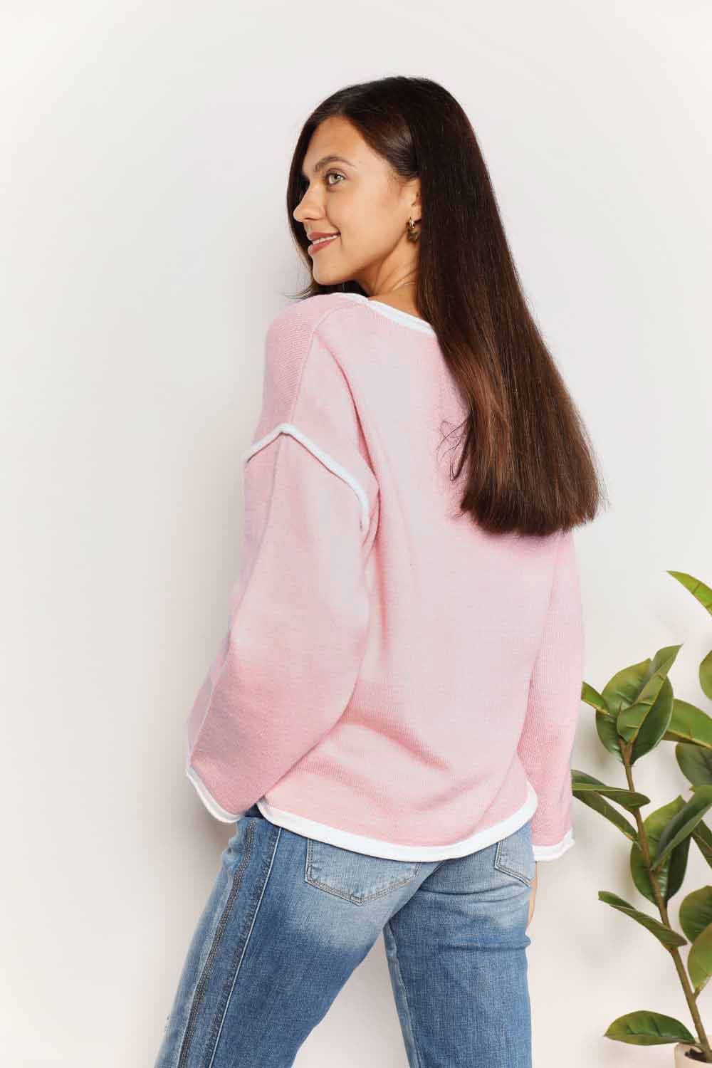 A woman with long curly hair is wearing the Angel Wings Contrast Detail Dropped Shoulder Knit Top in pink, paired with blue jeans, standing against a light background.