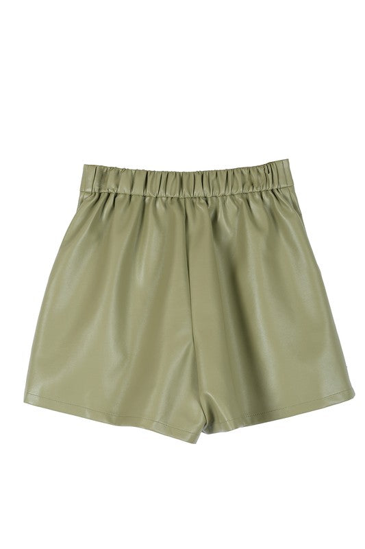 An individual is dressed in a light green sweater paired with pleated vegan leather shorts, featuring a functional pocket where one hand rests.