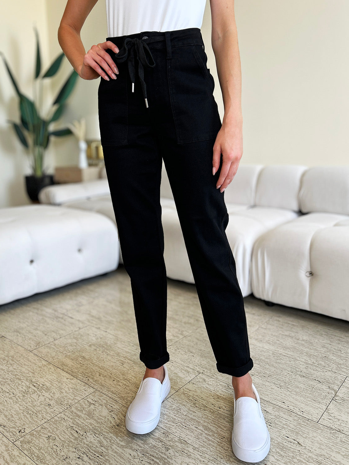 Person standing indoors, wearing a white top, black Judy Blue Full Size High Waist Double Roll Cuff Jeans, and white slip-on shoes, next to white couches and a plant in the background.