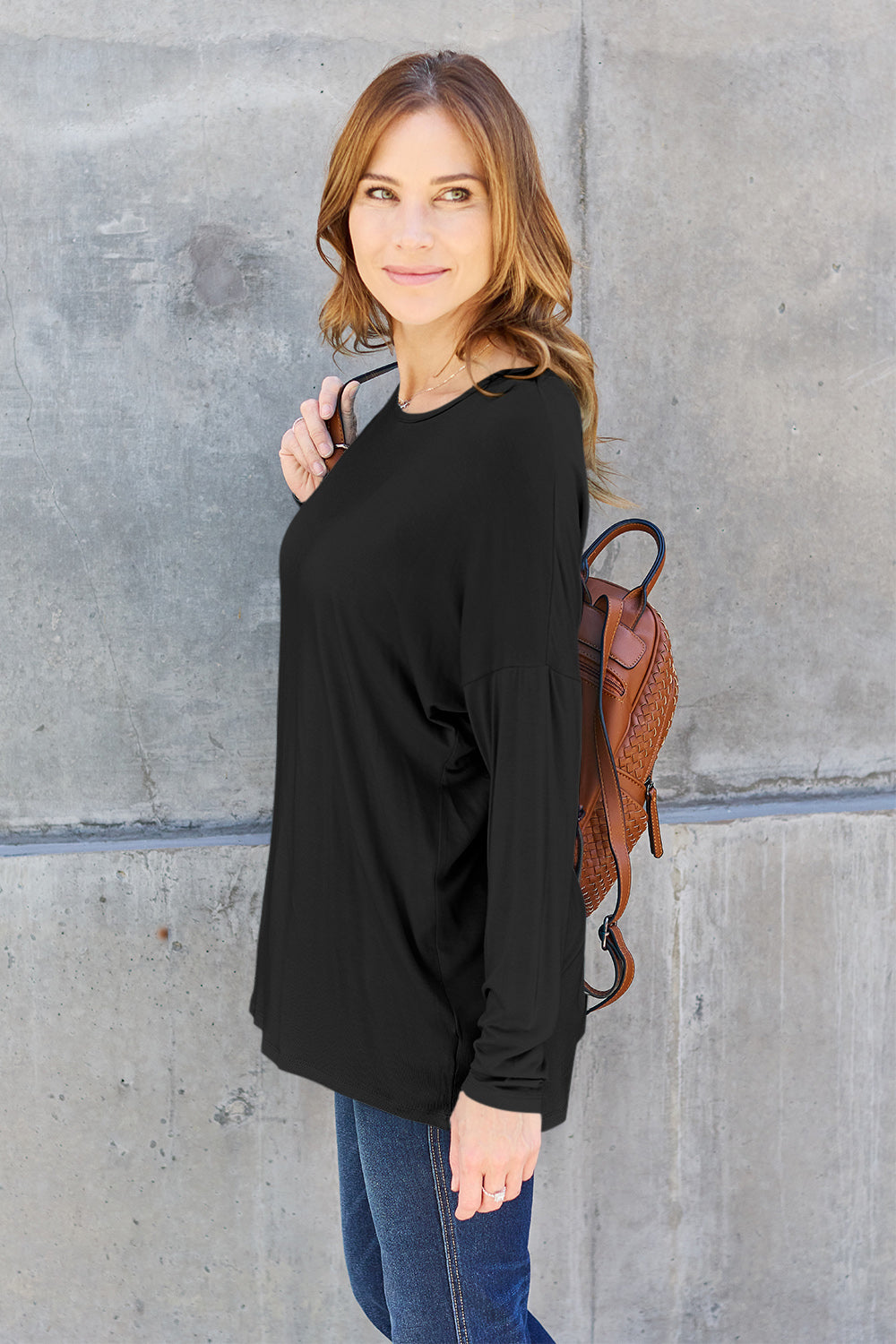 A woman with shoulder-length hair wearing a Basic Bae Full Size Round Neck Dropped Shoulder T-Shirt in green and a pair of jeans, exuding a basic style, stands against a concrete wall. She's holding an imported brown woven backpack on her shoulder while touching her hair with her hand.