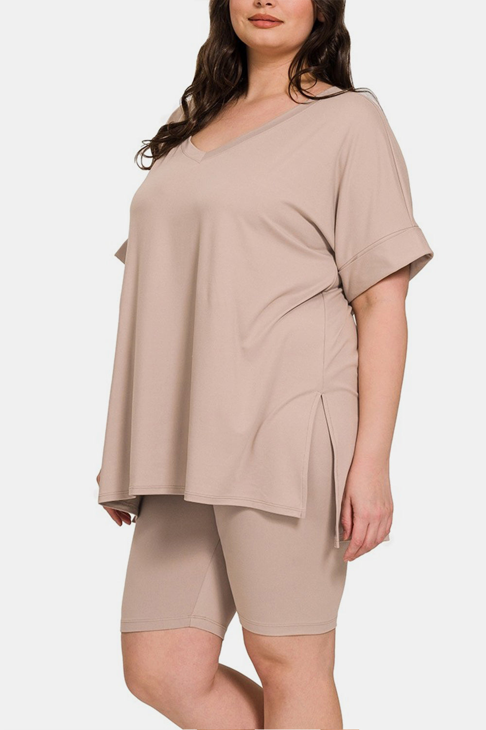 A woman wearing the highly stretchy Zenana Full Size V-Neck Short Sleeve Slit T-Shirt and Shorts Set in beige, featuring a mid-thigh shorts and short sleeve t-shirt combination, stands against a plain background.