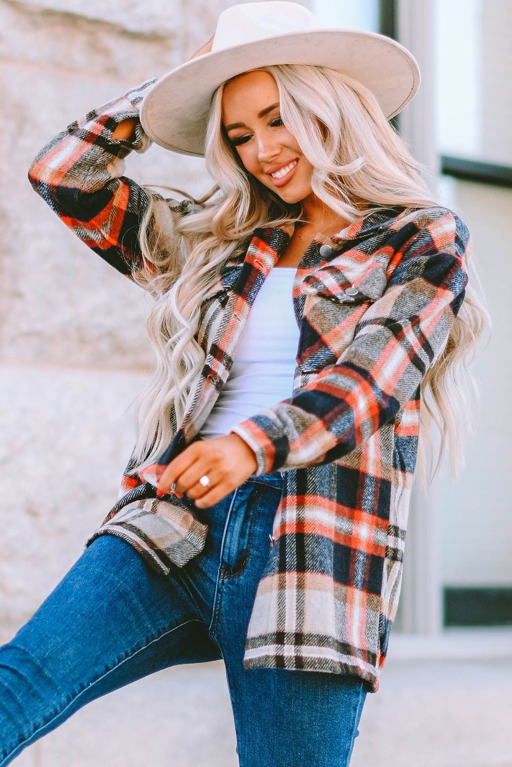 A person wearing a Brown Plaid Button Up Long Sleeve Flannel Shacket layered over a white top with ripped jeans and holding a brown handbag stands against a textured gray wall.